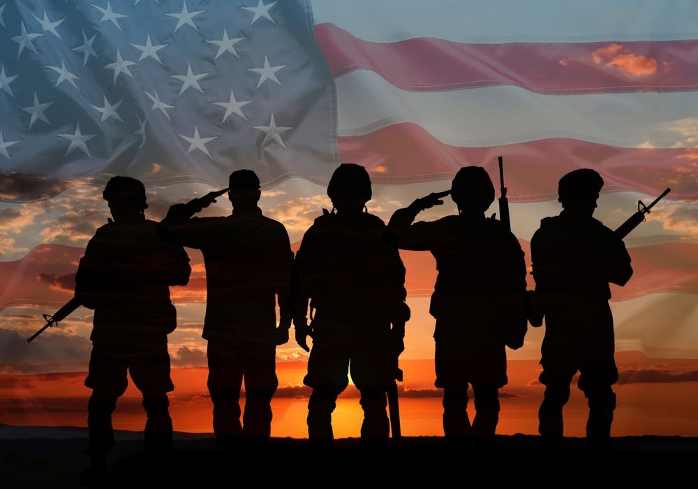 Silhouette of soldiers saluting against an American flag and sunset.