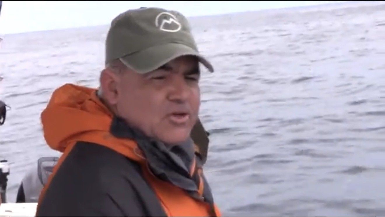 Man in orange jacket and green cap on a boat, speaking. Water and overcast sky in the background.