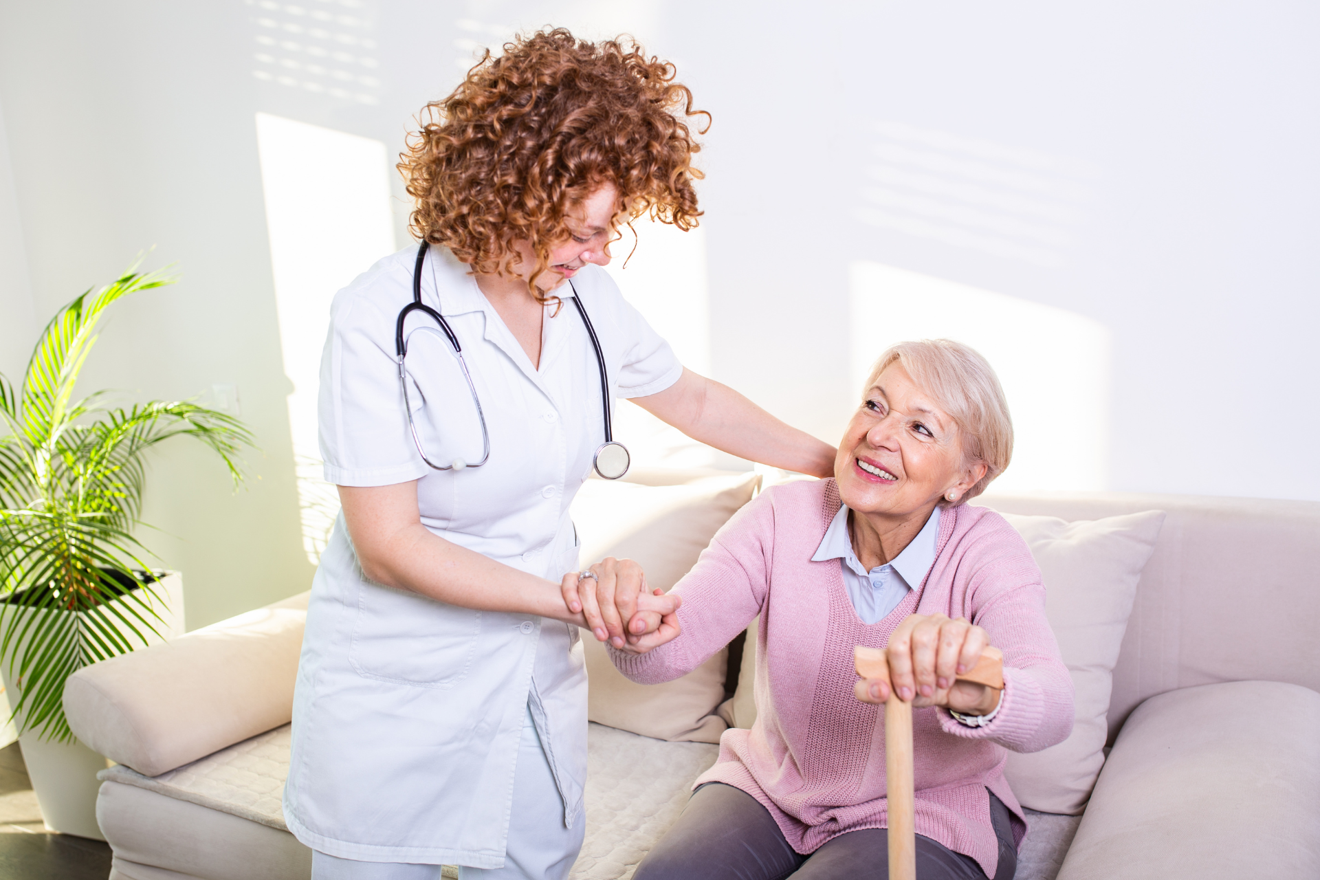 Nurse assisting an elderly patient with a cane on a sofa in a bright living room