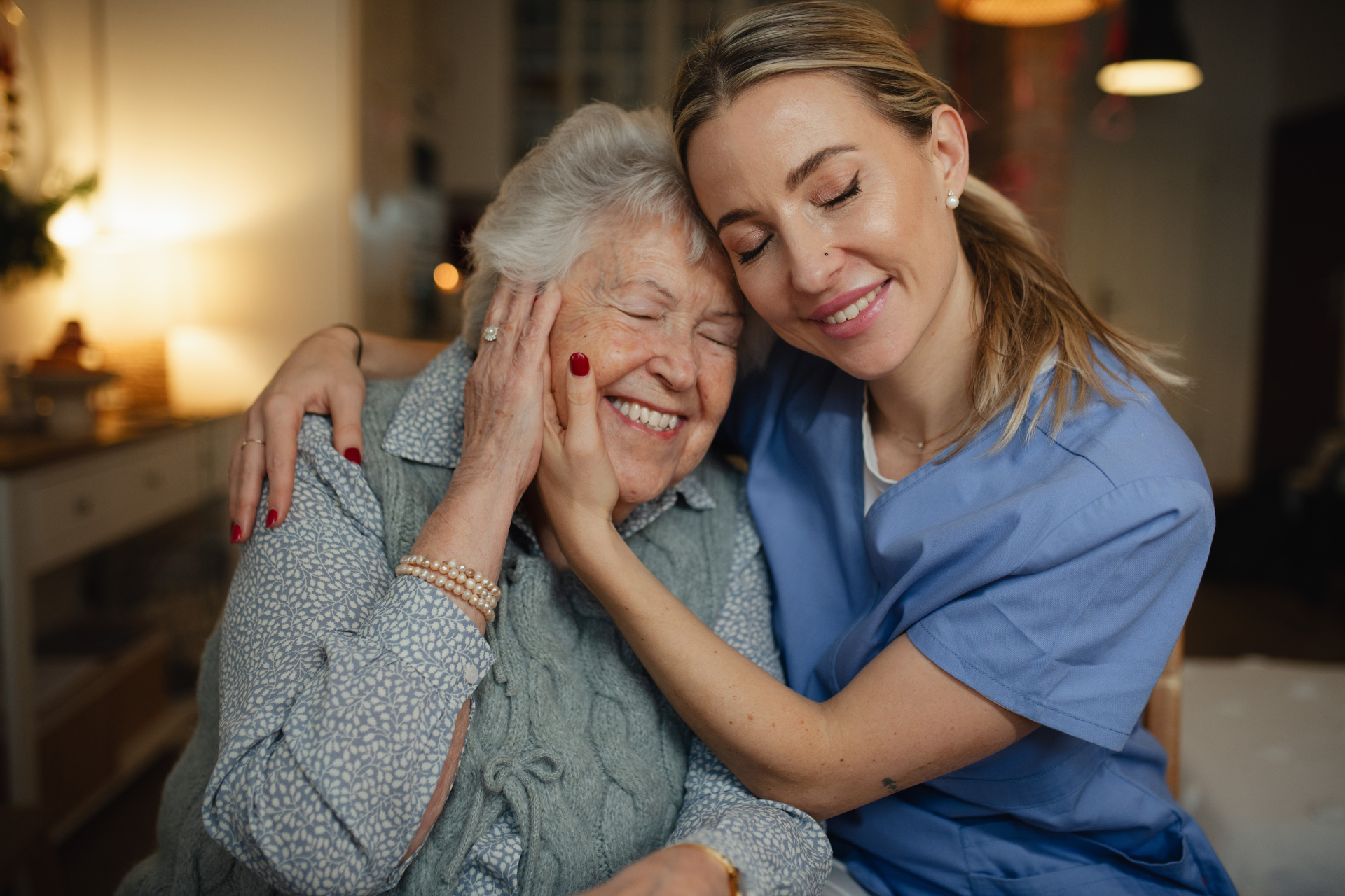 Older woman and younger caregiver embracing on a couch, both smiling in a warm home setting.