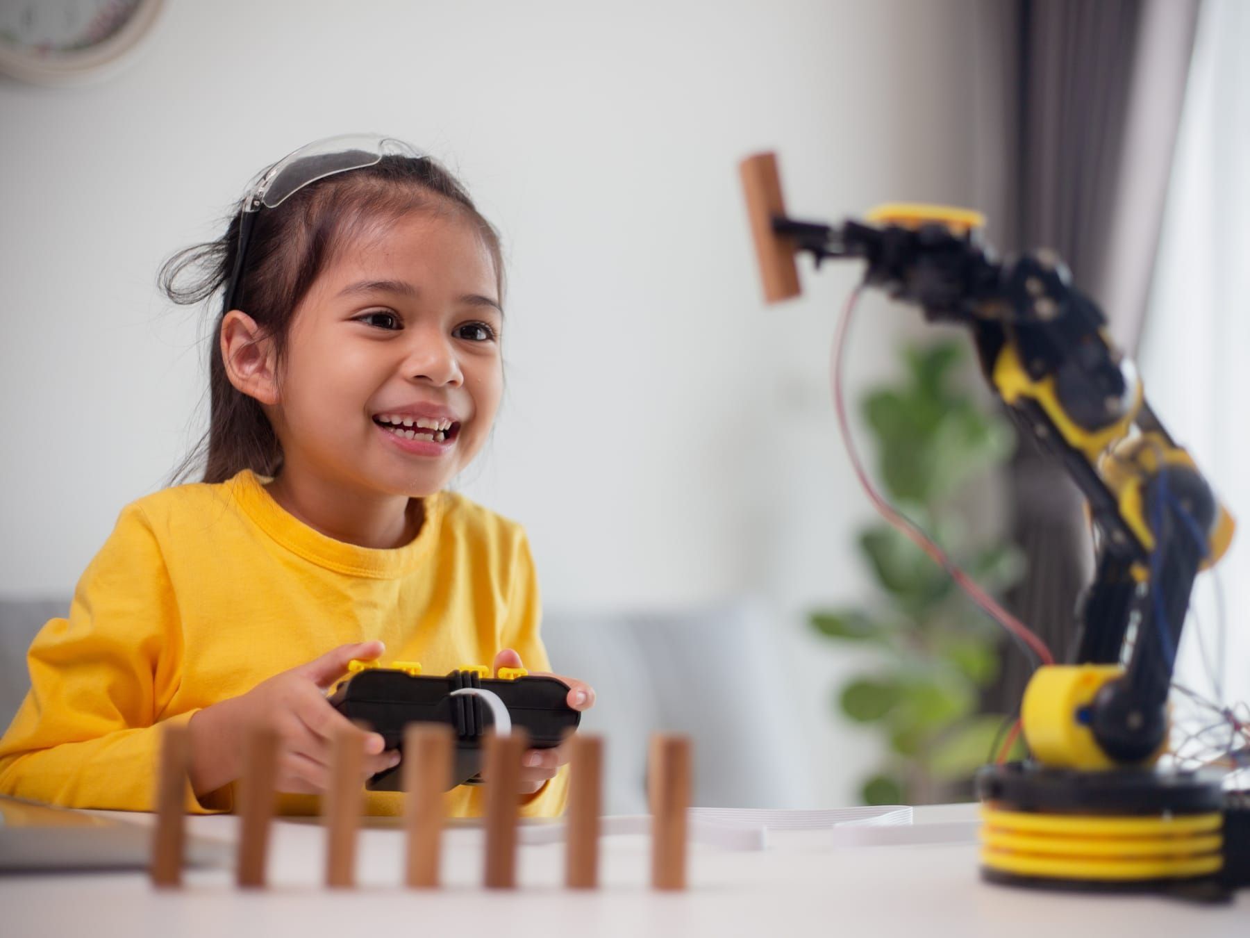 Girl in yellow operating a robotic arm, arranging wooden blocks on a table indoors.