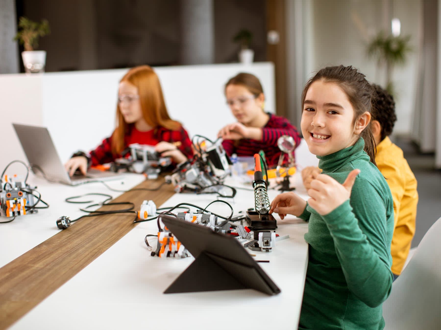 Girl in green turtleneck gives thumbs up at robotics class. Students use laptops and robots at a table.