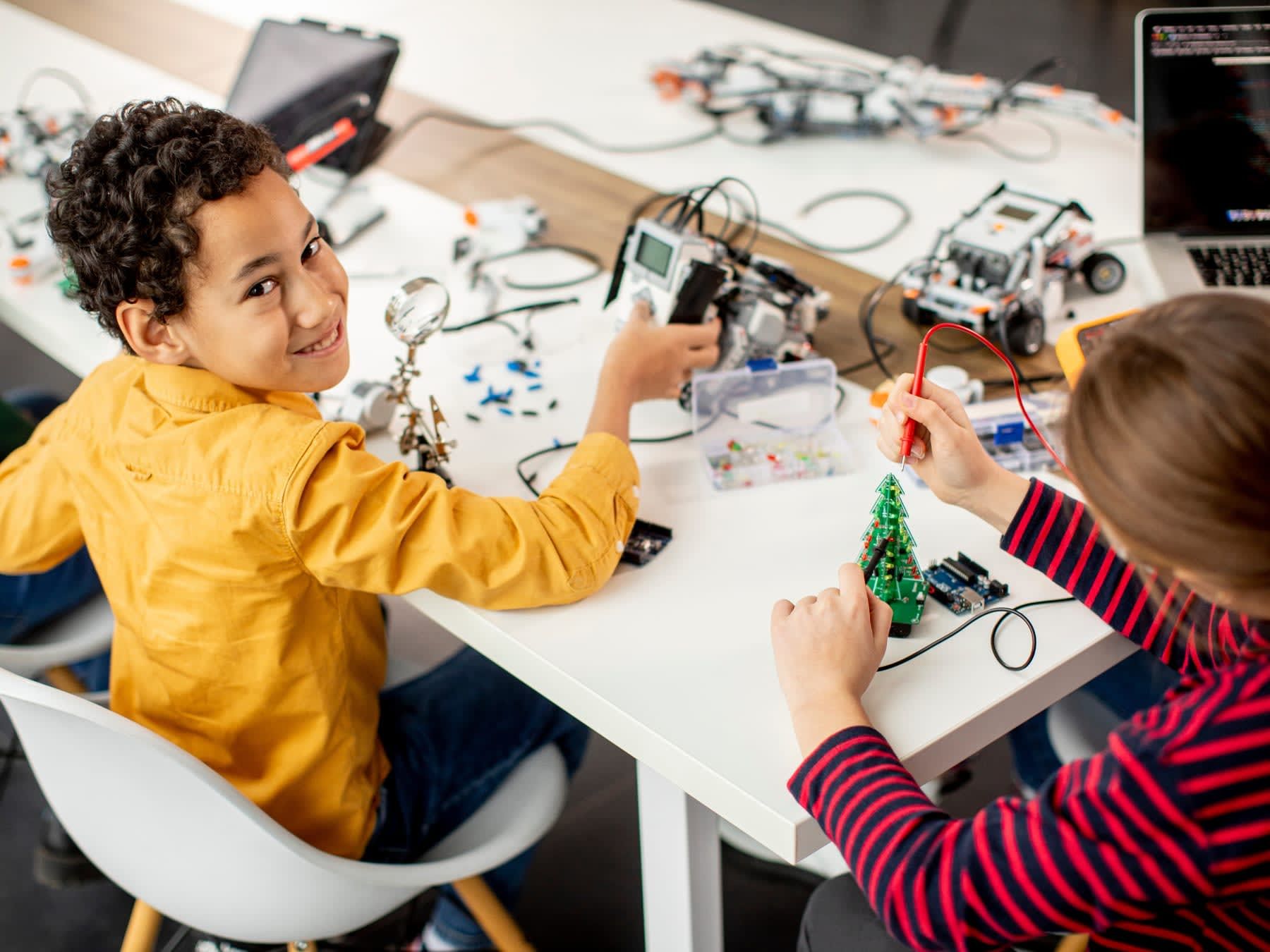 Boy and girl building robots at a table, smiling. Yellow shirt, red striped shirt, robotics lab setting.
