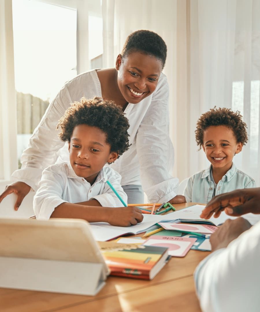 Woman smiling, helping two young boys with homework at a table indoors. Bright, sunny room.