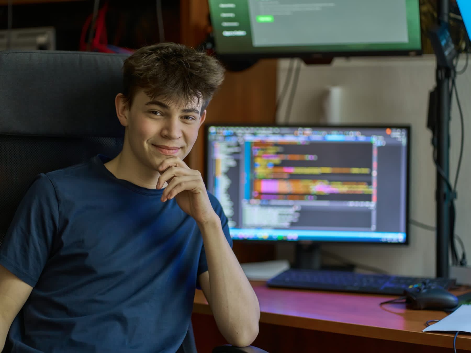 Young man smiling, sitting at desk with computer screens displaying code.