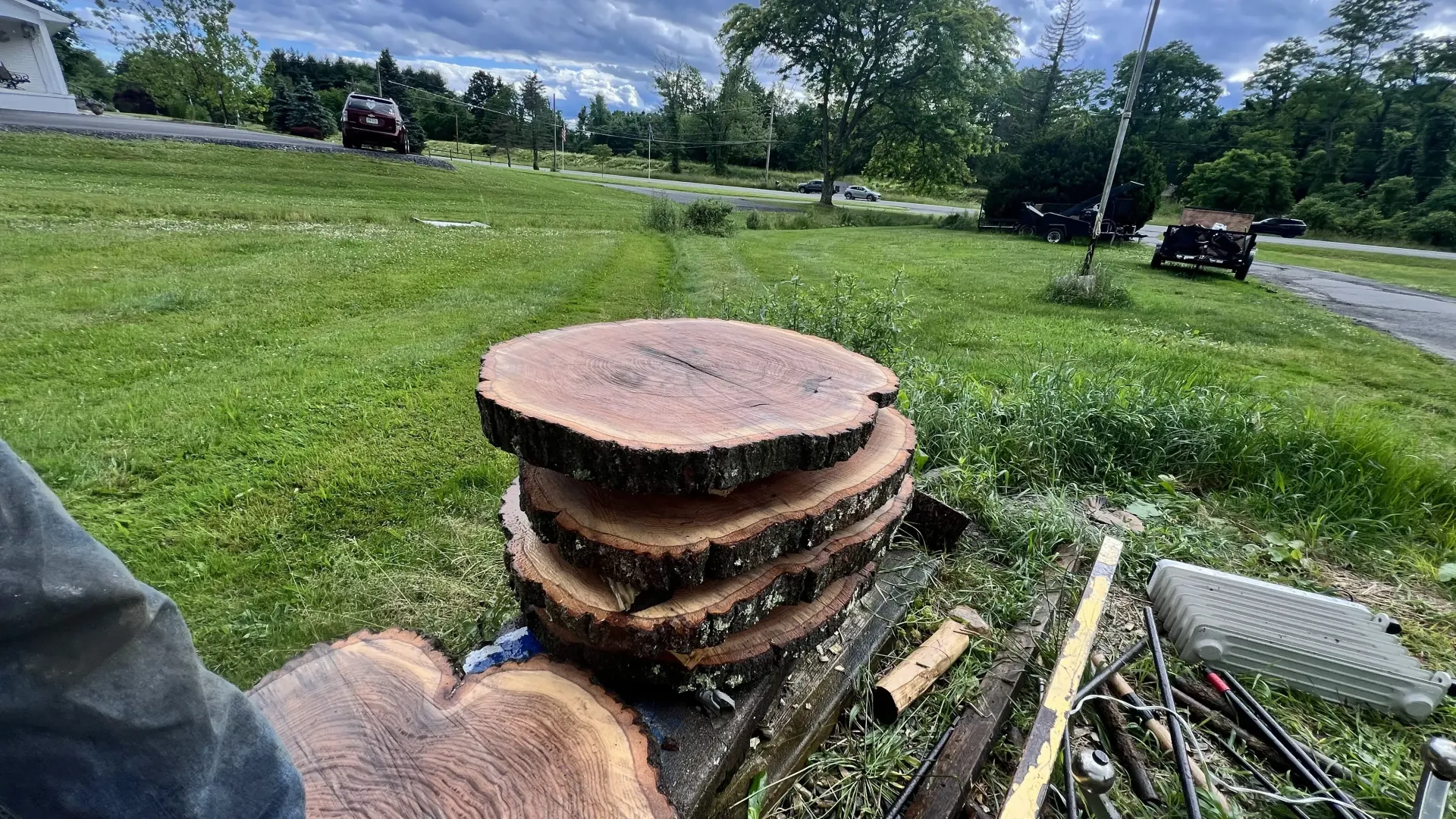 Stacked wooden tree stumps in a grassy yard near a road, with tools and logs nearby.