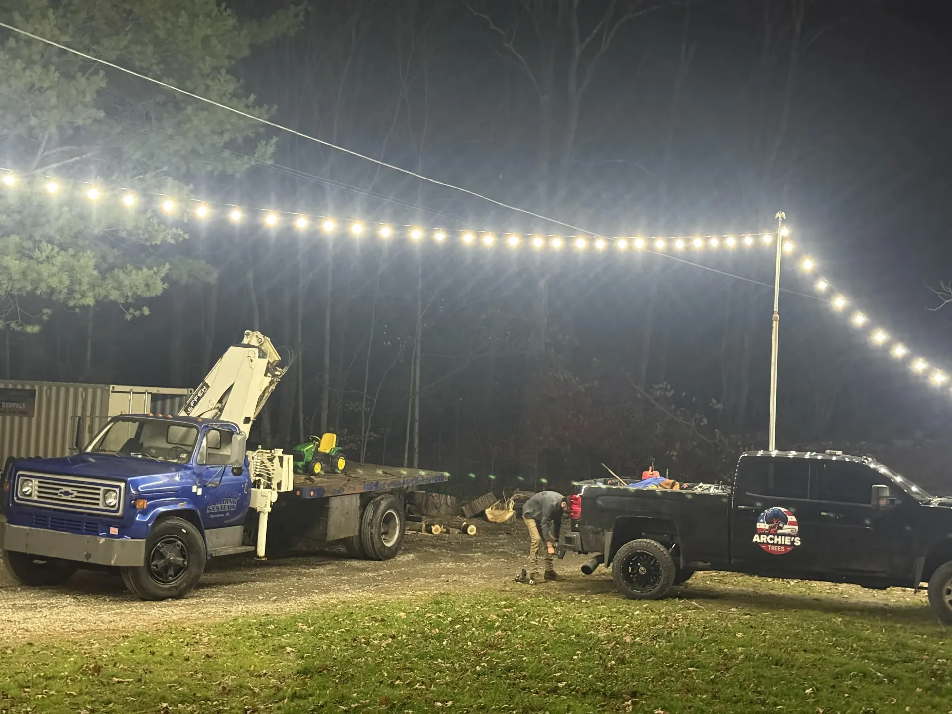 Tow trucks and pickup trucks parked at night under string lights in a wooded area