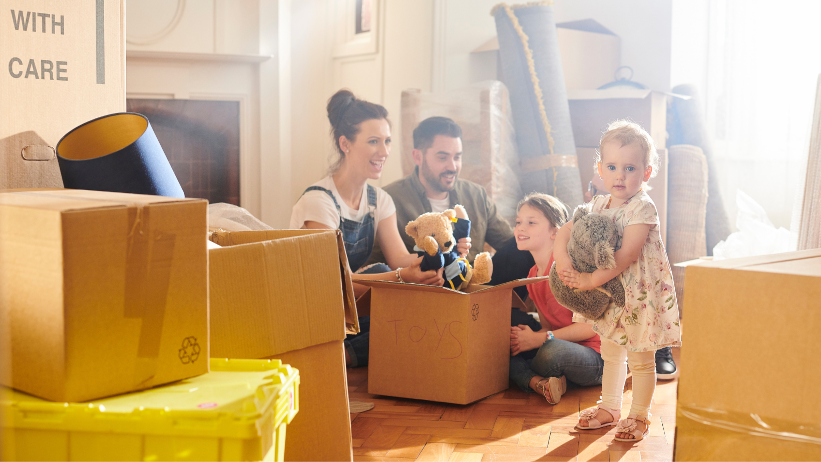 A family is sitting on the floor in a living room surrounded by cardboard boxes.