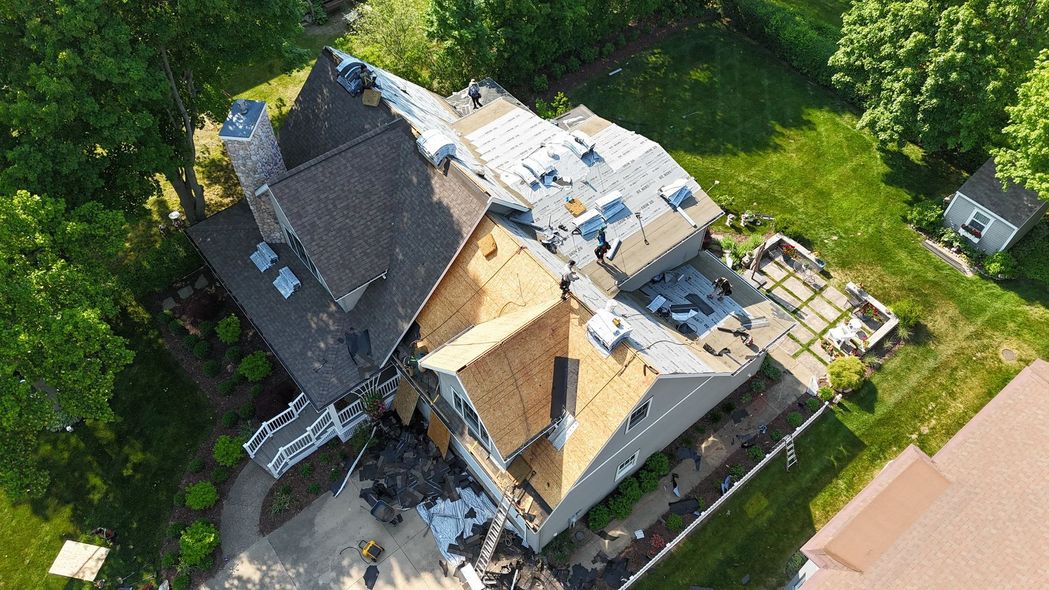 Aerial view of a gray house with a damaged roof beside lush green trees and neighboring homes.