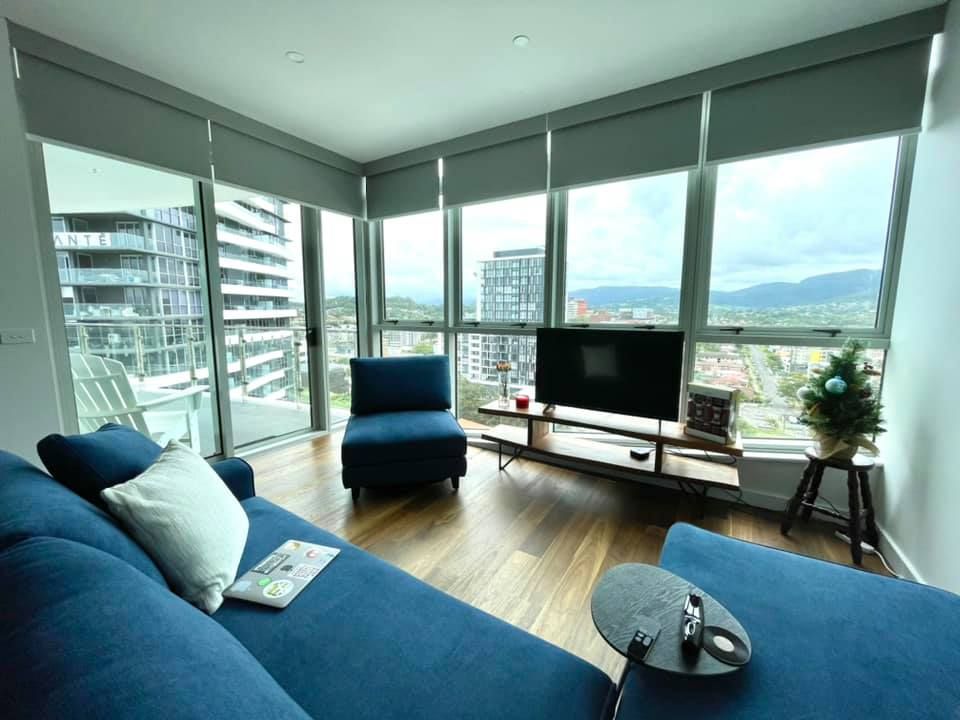A modern living room with a blue sectional sofa, a single blue armchair, and large windows showcasing an outdoor view. — Glenn Koek - Shutters - Curtains - Blinds in West Wollongong, NSW