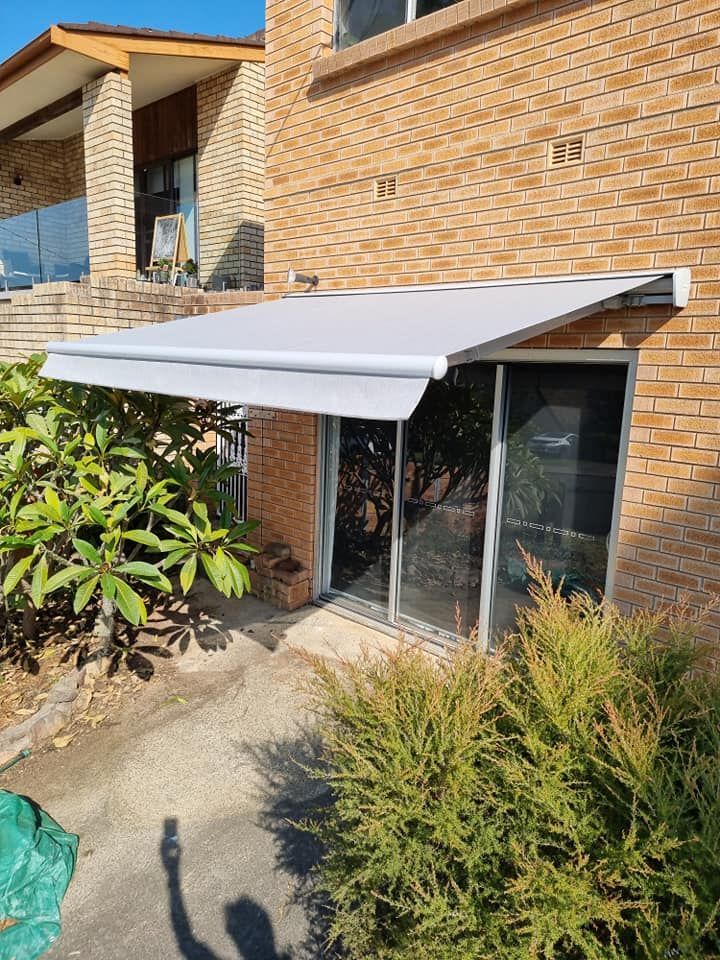 A white retractable awning over glass sliding doors on a brick building, with lush green bushes in front. — Glenn Koek - Shutters - Curtains - Blinds in West Wollongong, NSW