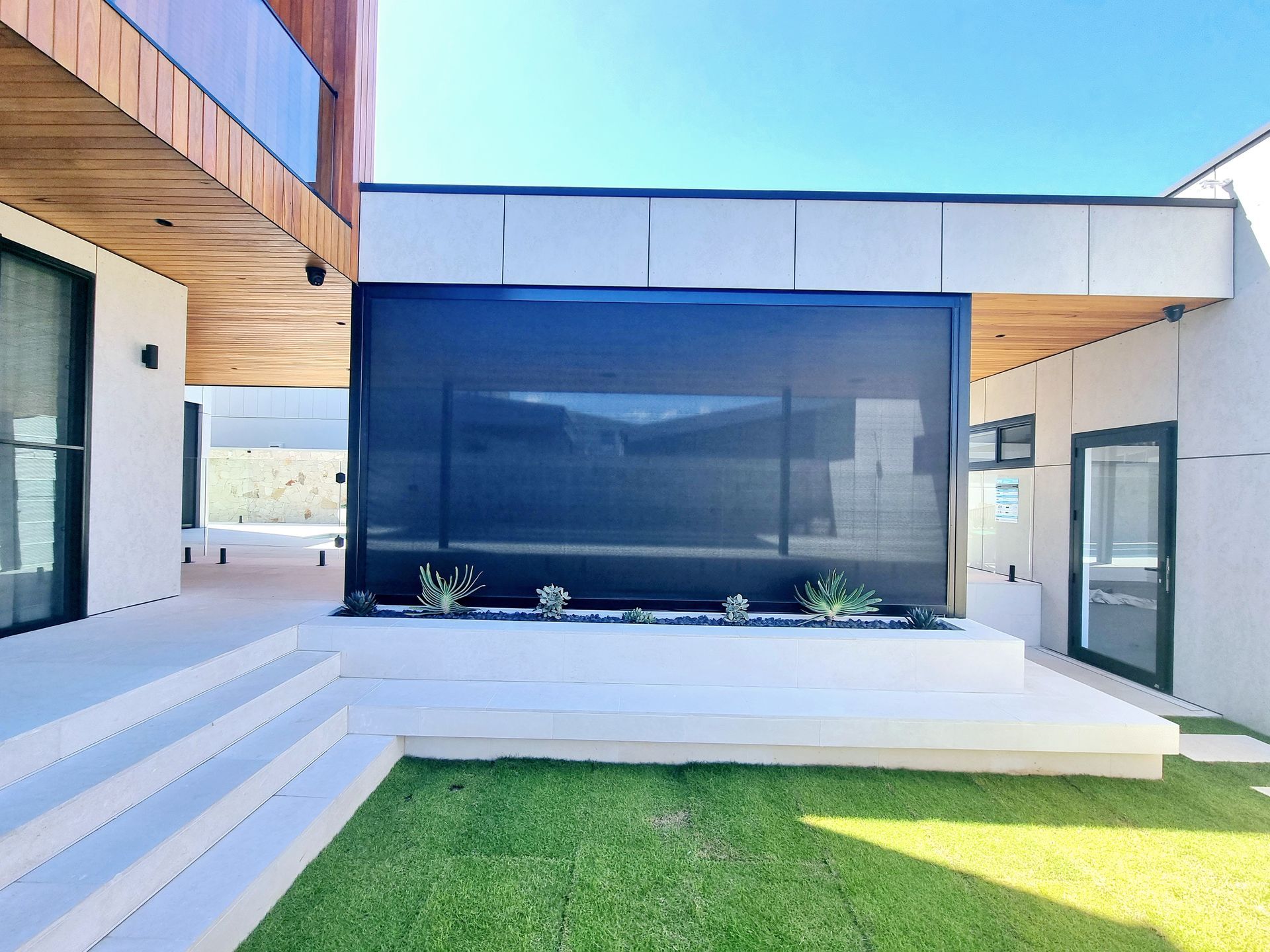 Modern home exterior with a large retractable black shade over a raised patio, white steps, and lawn. — Glenn Koek - Shutters - Curtains - Blinds in West Wollongong, NSW