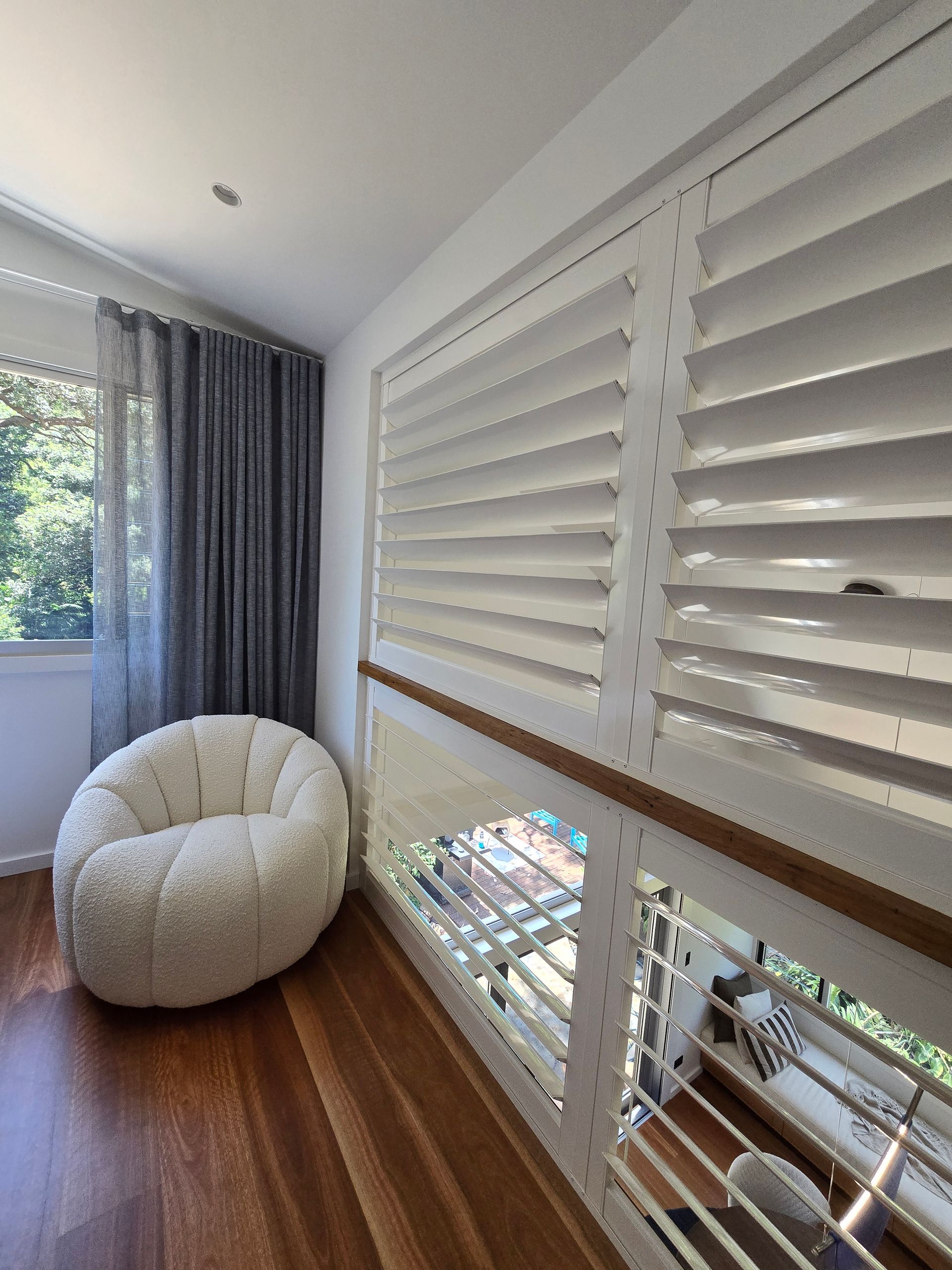 Cozy room with white shutters, wooden floors, and a fuzzy white chair near a window with gray curtains. — Glenn Koek - Shutters - Curtains - Blinds in Sutherland Shire, NSW
