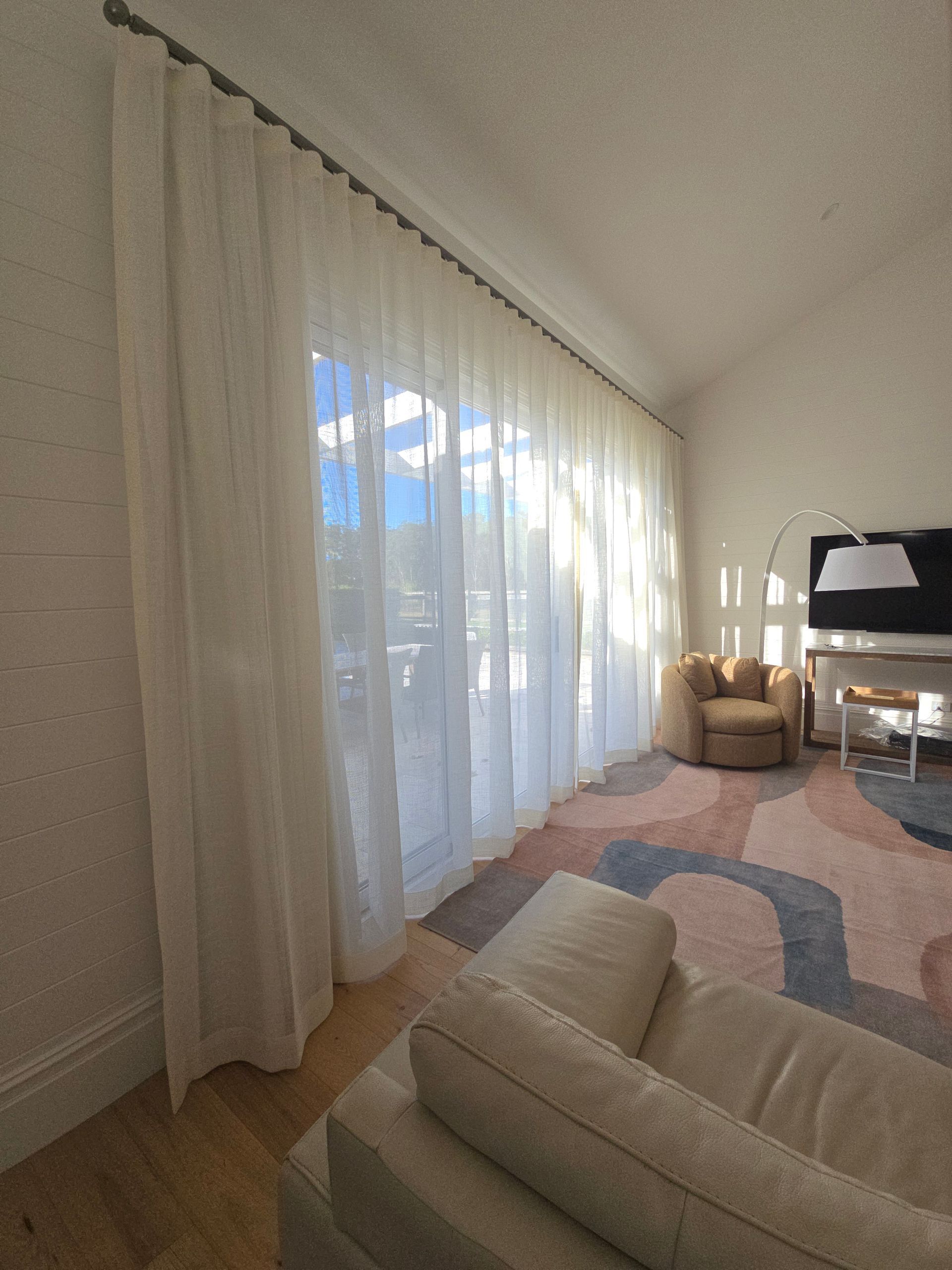 Sheer white curtains frame a bright window in a living room with a patterned rug and cream-coloured sofa. — Glenn Koek - Shutters - Curtains - Blinds in West Wollongong, NSW