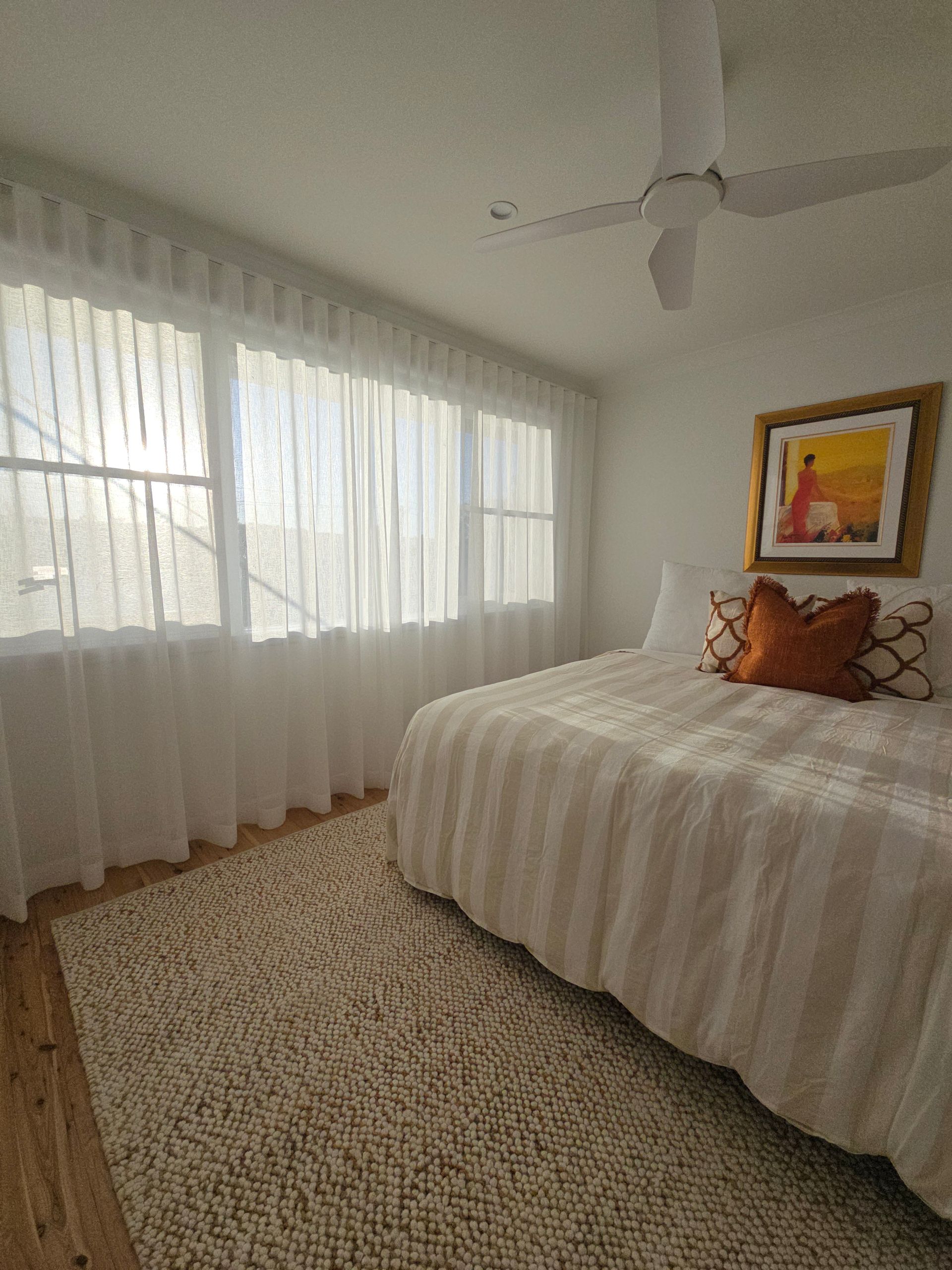 A bedroom with white walls, sheer curtains, a bed, area rug, and artwork. — Glenn Koek - Shutters - Curtains - Blinds in West Wollongong, NSW