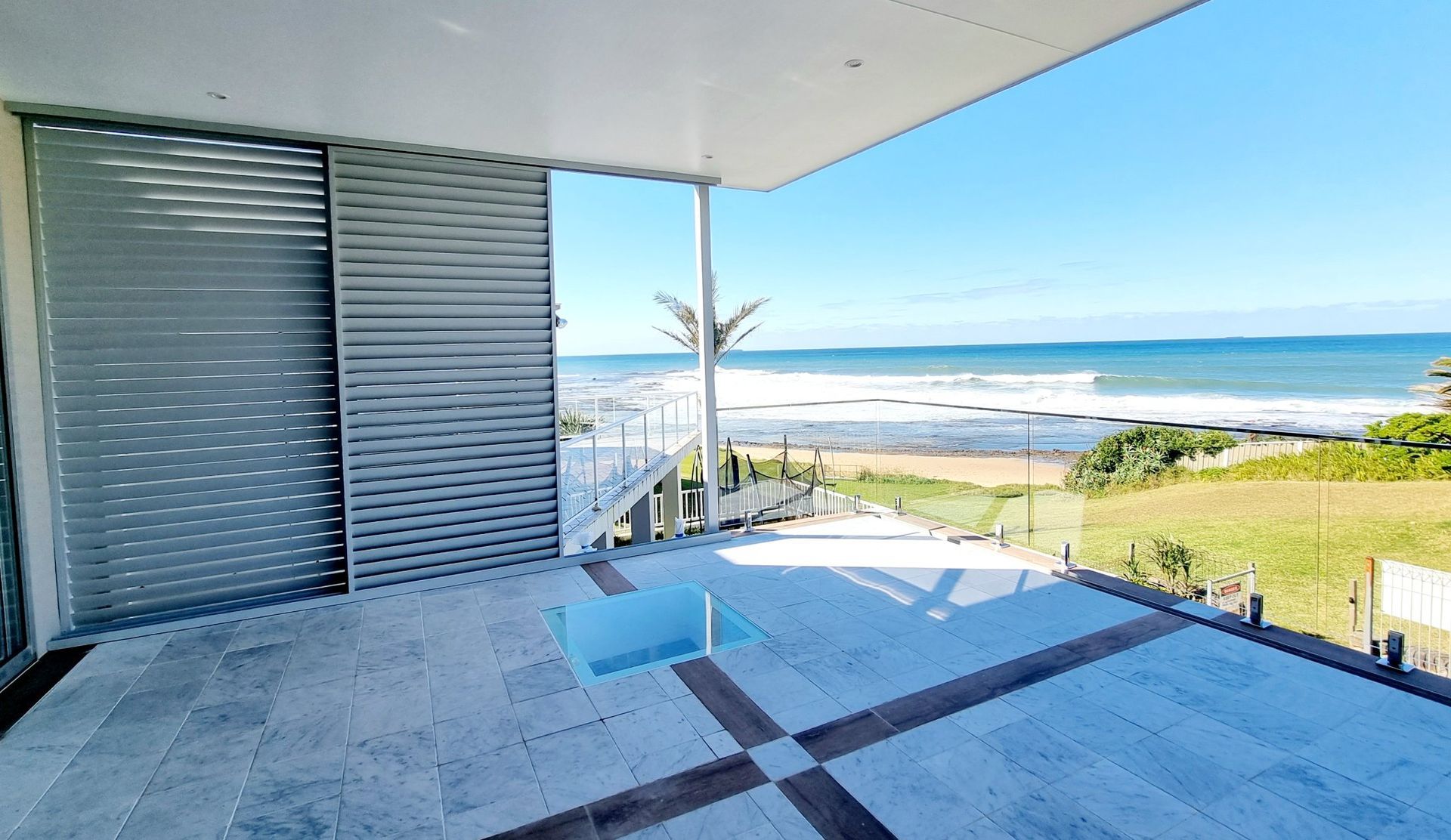 Oceanfront balcony with a view of the beach and ocean. Sliding door, tiled floor, and blue sky. — Glenn Koek - Shutters - Curtains - Blinds in West Wollongong, NSW