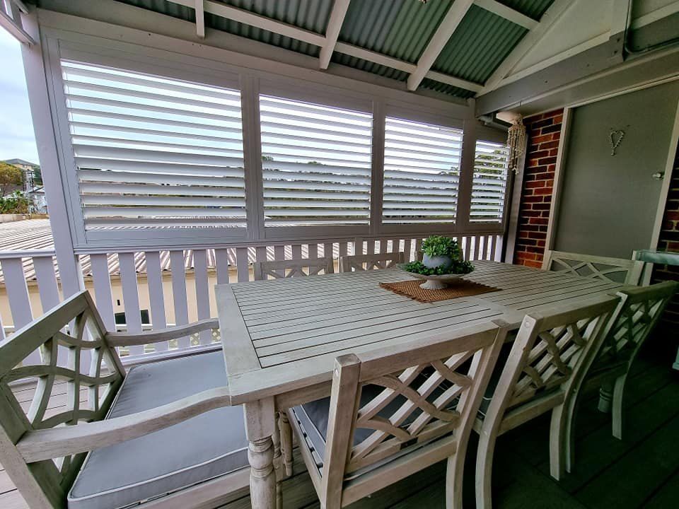 Outdoor dining area with white table, chairs, and slatted shutters. — Glenn Koek - Shutters - Curtains - Blinds in West Wollongong, NSW