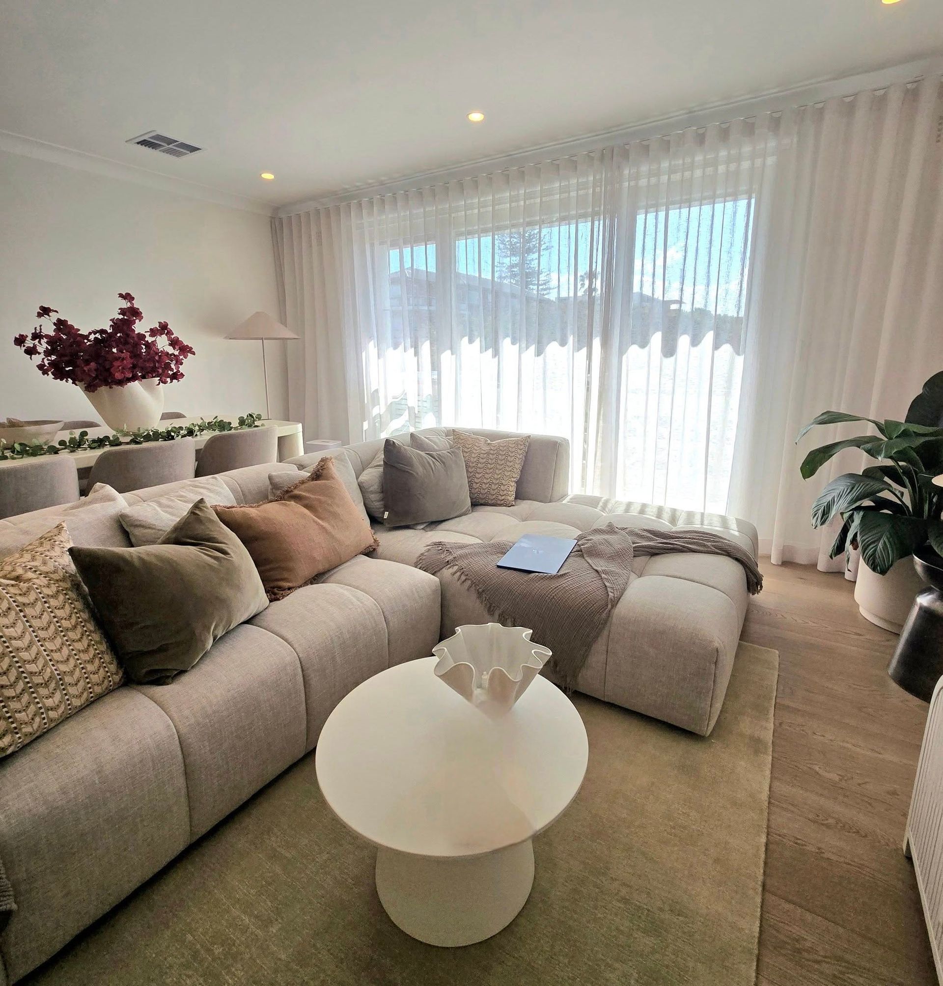 A modern living room with a beige sectional sofa, coffee table, and sheer white curtains. Natural light streams in. — Glenn Koek - Shutters - Curtains - Blinds in West Wollongong, NSW