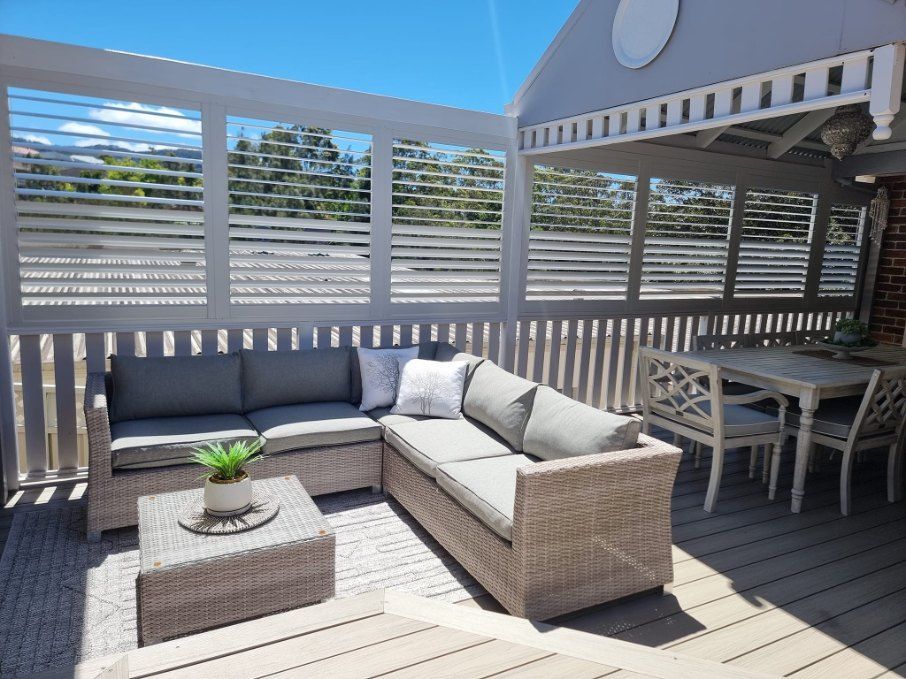 Outdoor patio with a gray sectional sofa, table, and dining table with chairs. Wooden deck and shutter wall. — Glenn Koek - Shutters - Curtains - Blinds in West Wollongong, NSW