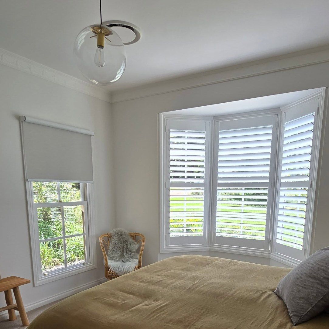 Bedroom with white walls, a bay window with white shutters, a bed with a gold-coloured cover, and a small stool. A hanging light fixture is visible. — Glenn Koek - Shutters - Curtains - Blinds in West Wollongong, NSW