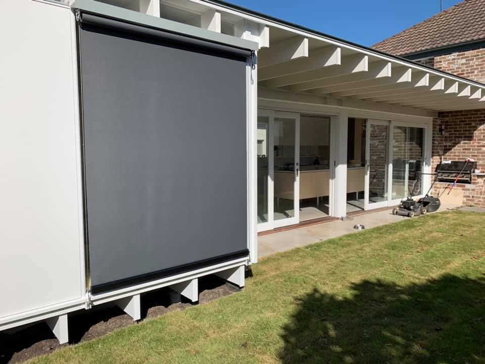 A grey sunshade covers part of a white building next to a grassy yard. Sliding glass doors and a brick building are in the background. — Glenn Koek - Shutters - Curtains - Blinds in West Wollongong, NSW