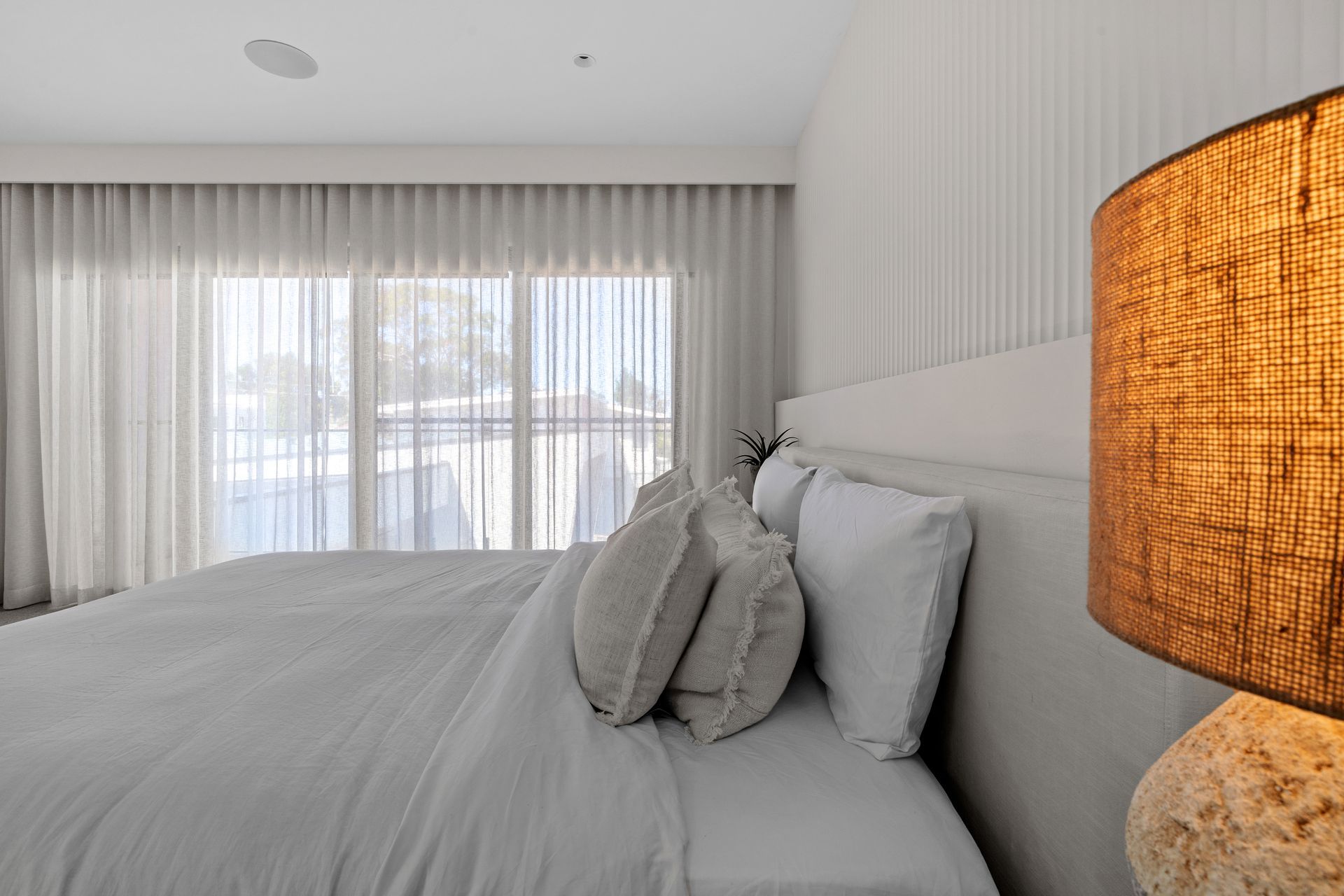 Bedroom with bed, white linens, pillows, sheer curtains, and a lit lamp. — Glenn Koek - Shutters - Curtains - Blinds in Sutherland Shire, NSW