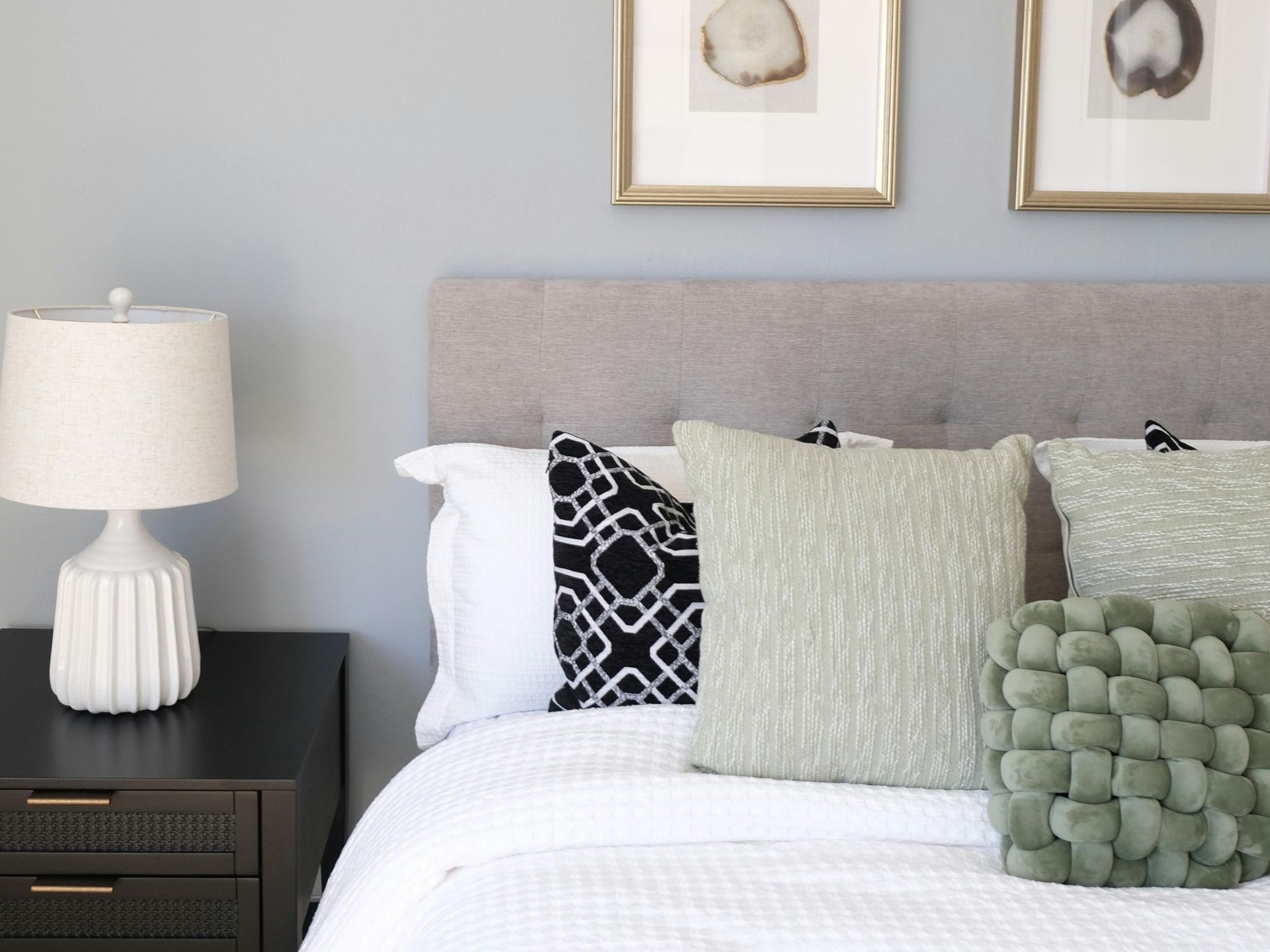 A modern bedroom with a grey tufted headboard, white bedding, accent pillows, a white lamp, and framed wall art.