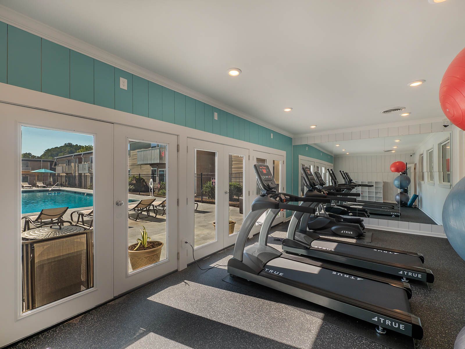 A gym with several treadmills lined up along a wall of glass doors that open to an outdoor swimming pool.