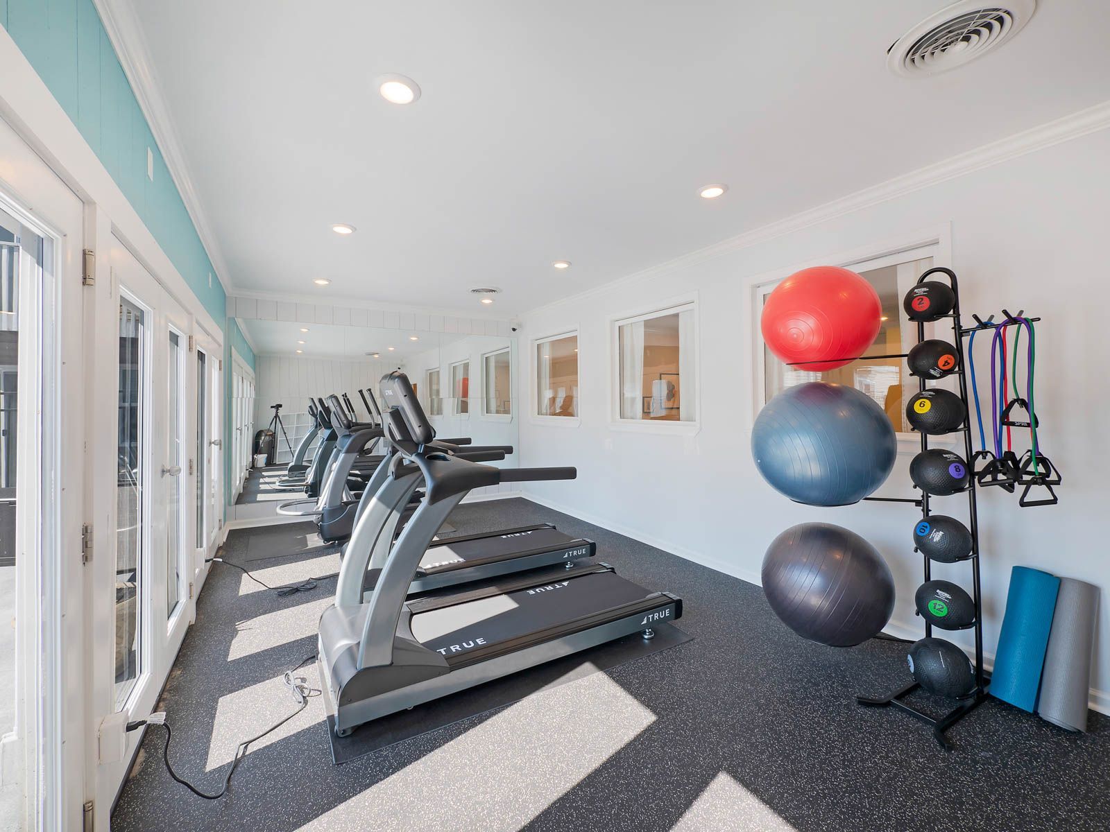 A bright, clean gym with rows of treadmills, exercise balls on a rack, and yoga mats on a dark, speckled floor.