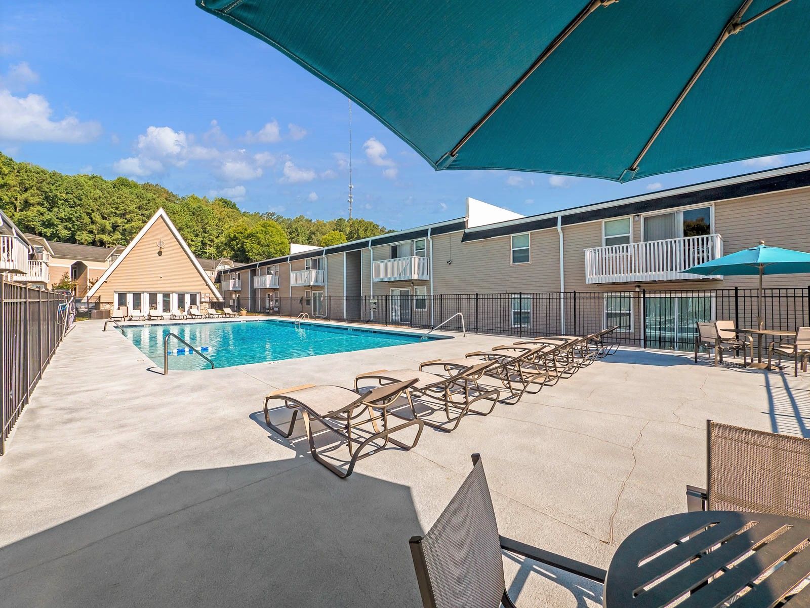 An outdoor swimming pool surrounded by a concrete deck with lounge chairs and tables under a teal umbrella.