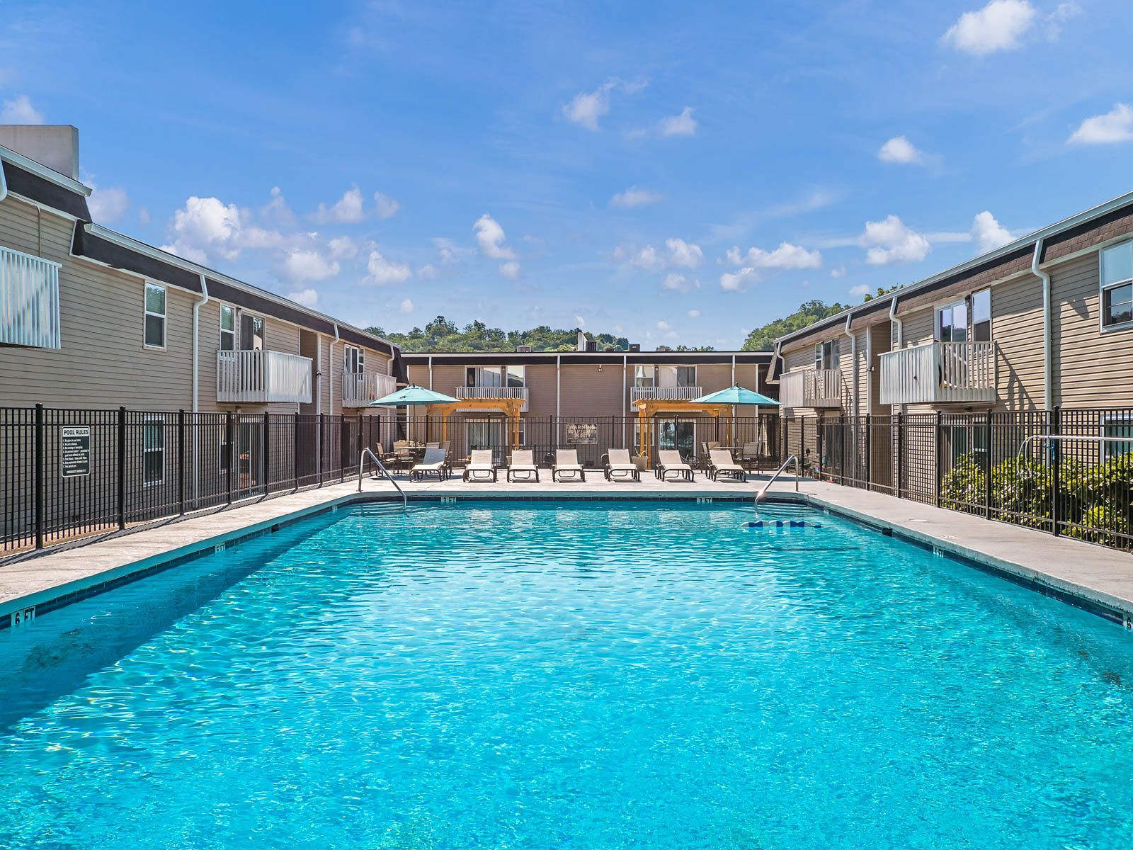 A bright blue swimming pool centered between two apartment buildings with deck chairs and umbrellas on the patio.