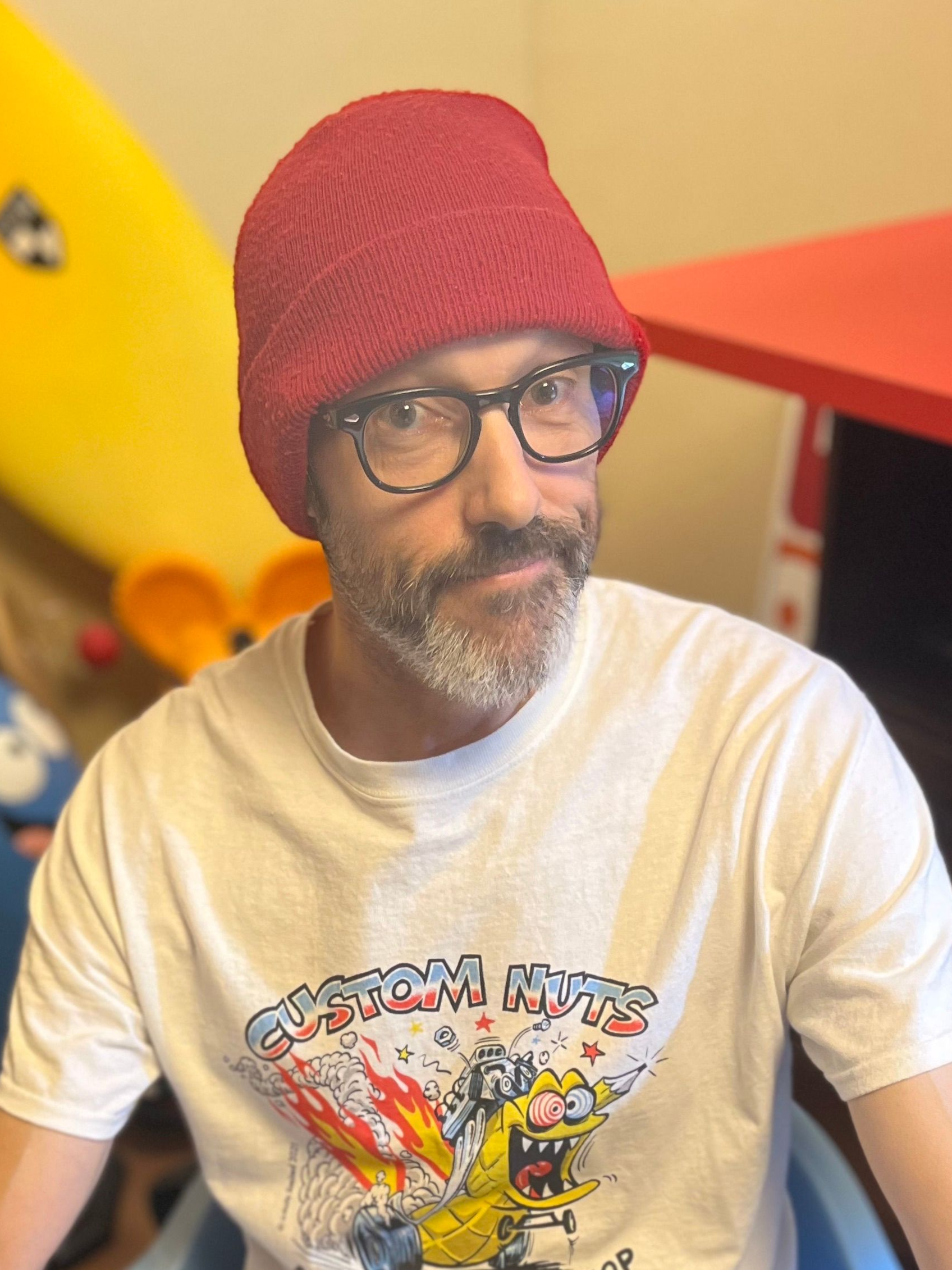 A person with a beard, wearing a red beanie, glasses, and a graphic t-shirt, sits in a room with a yellow surfboard.