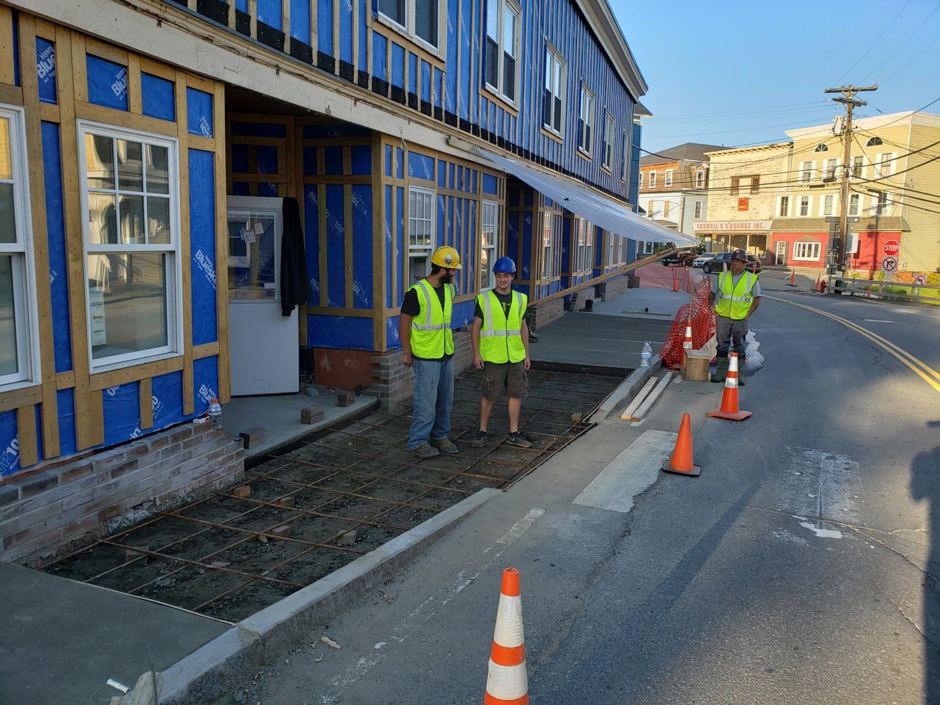 Construction workers in neon vests repairing a sidewalk beside a blue building on a city street.