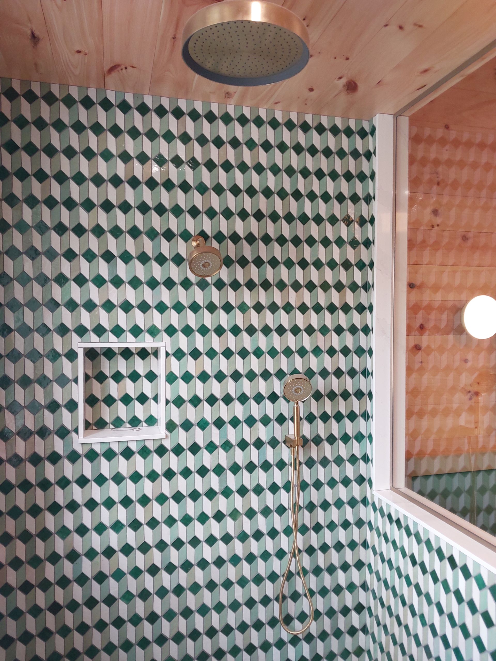 Small tiled shower with green-and-white pattern, overhead showerhead, and wooden ceiling.