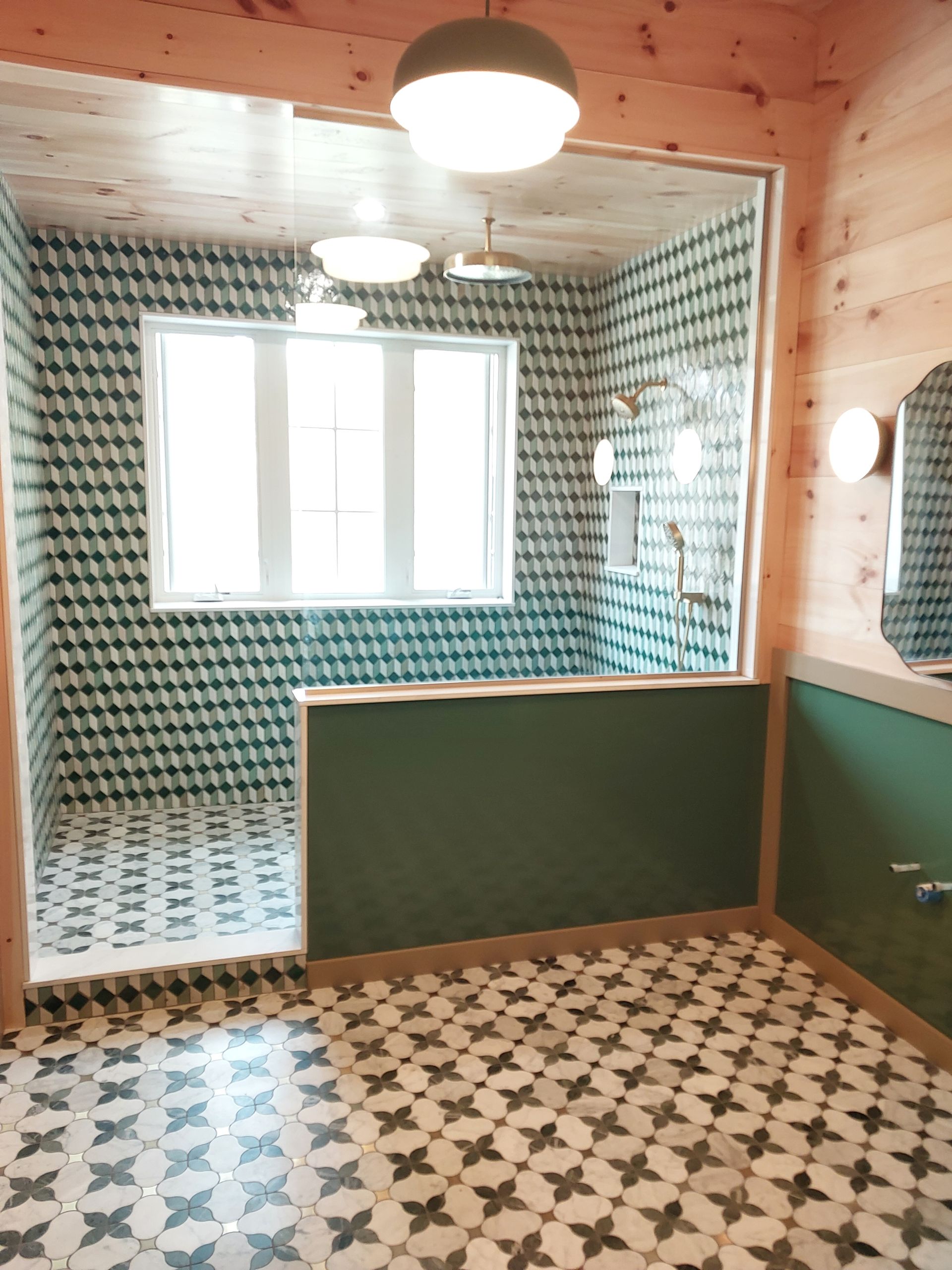 Small tiled bathroom with green half-walls, patterned floor, wood-paneled ceiling, and a window.
