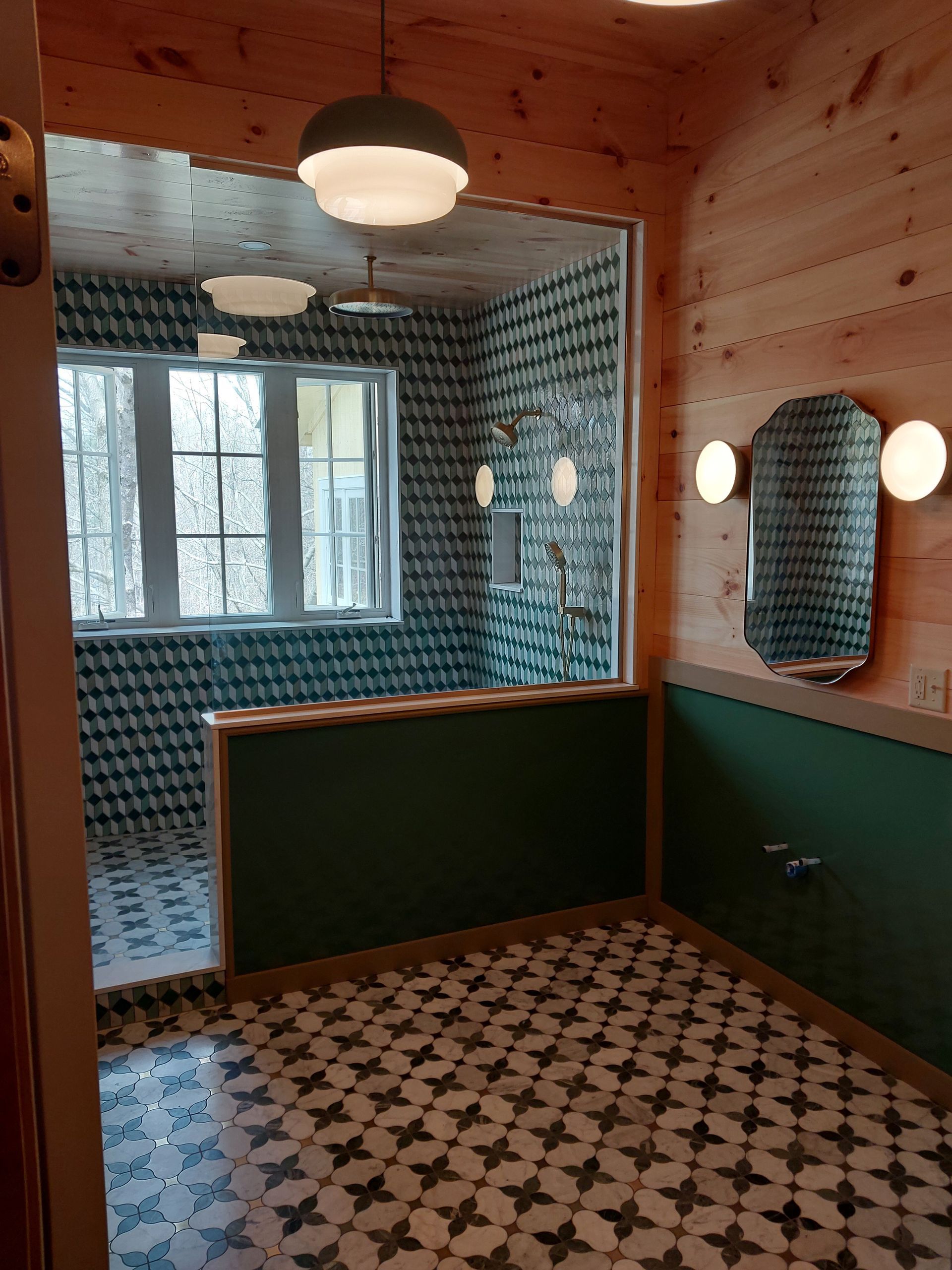 Small bathroom with blue patterned tile, wood walls, a window, round mirror, and patterned floor.