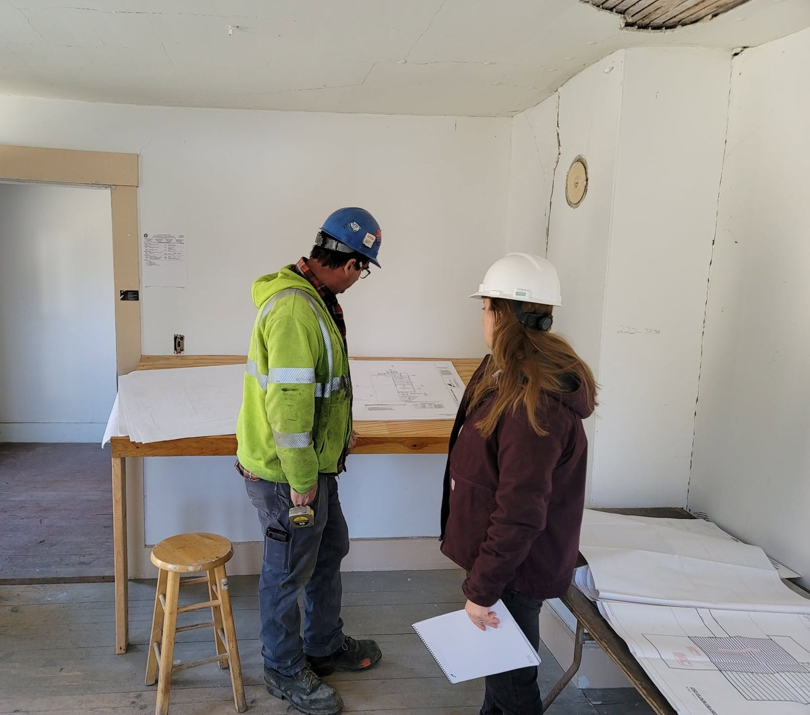 Two workers in hard hats discuss plans inside a bare room under renovation.