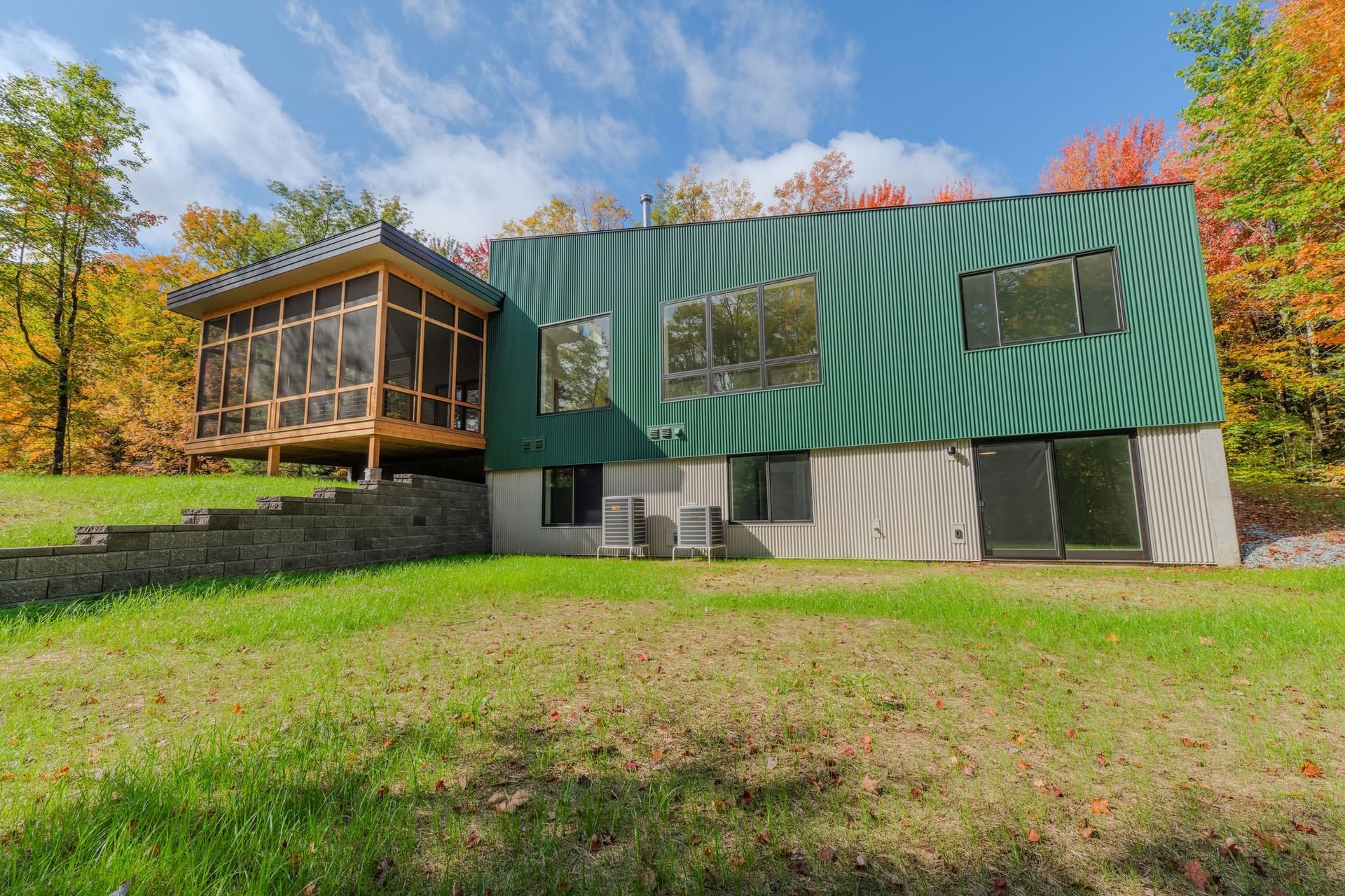 Modern house with green metal siding and a wooden deck on a grassy hillside in autumn