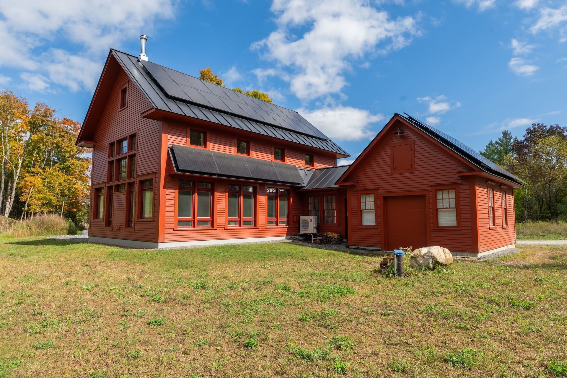Red wooden house and barn on a grassy field under a blue sky with autumn trees