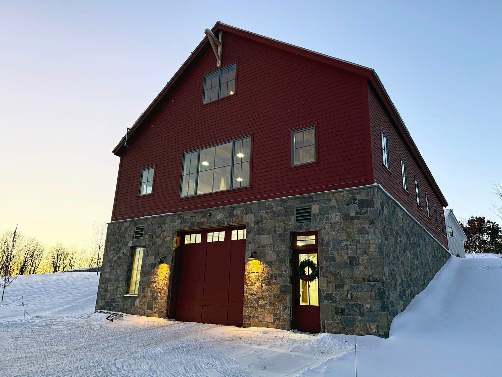 Red barn-style building with stone lower level and lit doorway in a snowy landscape at dusk