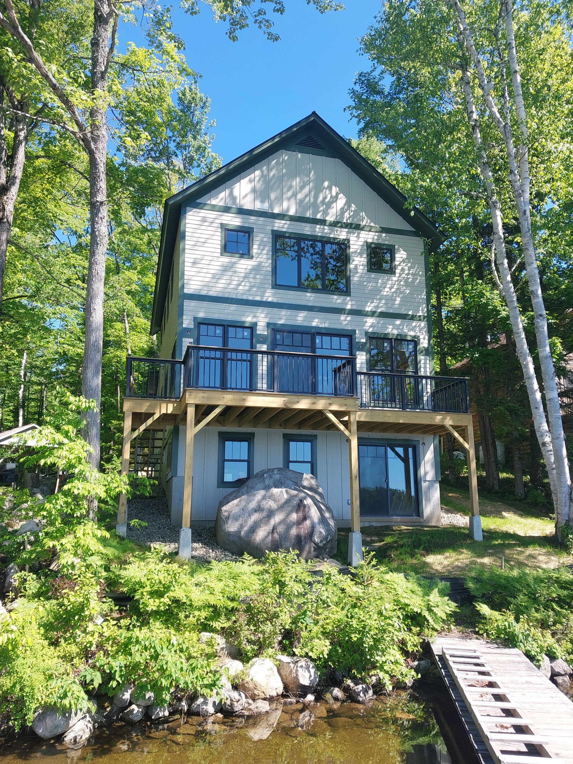White two-story cabin with blue trim on a wooded lakeshore, with a dock in the foreground.