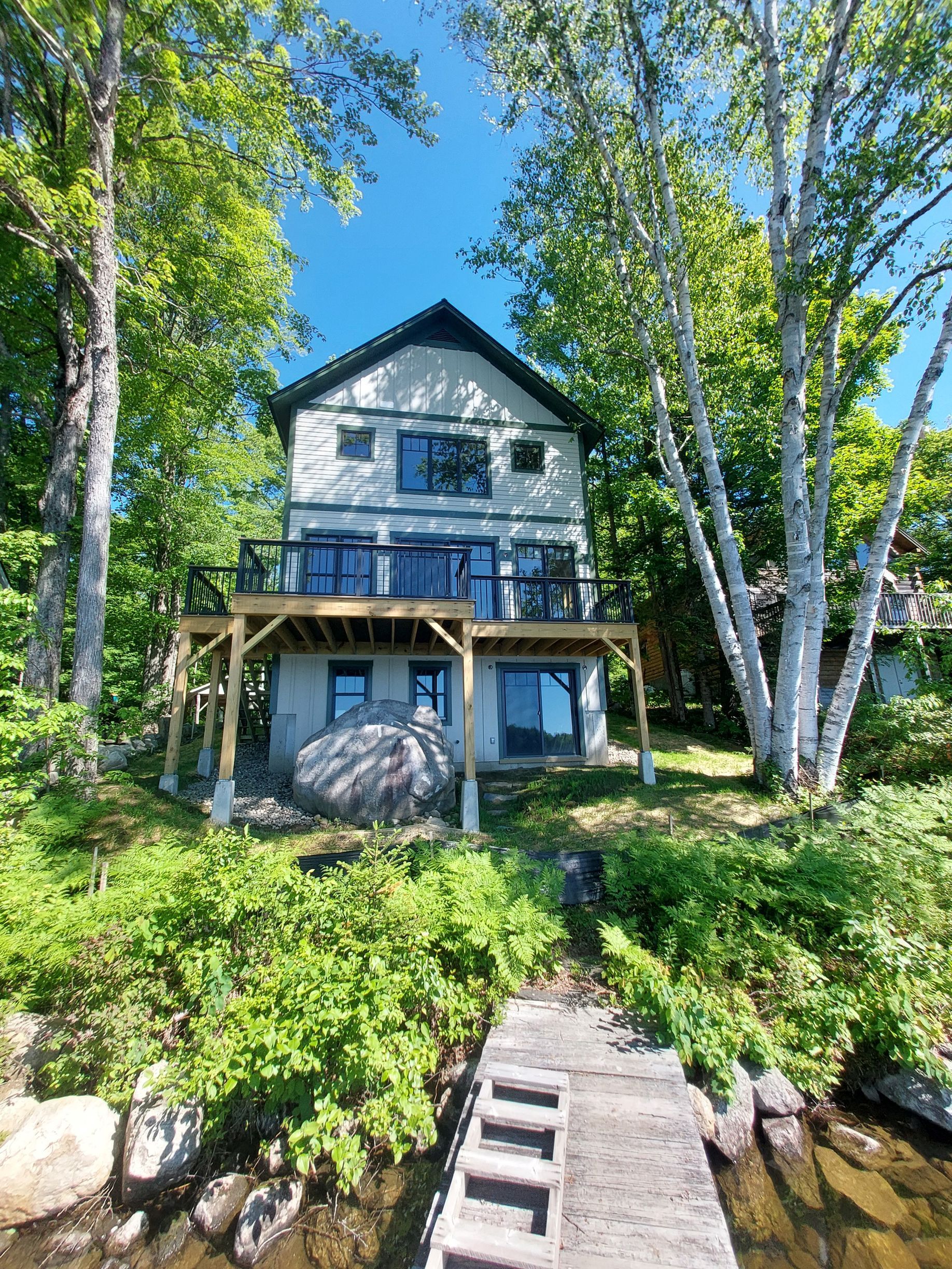 Blue lakeside cabin among tall birch trees, with a wooden deck and stone steps leading to the water.