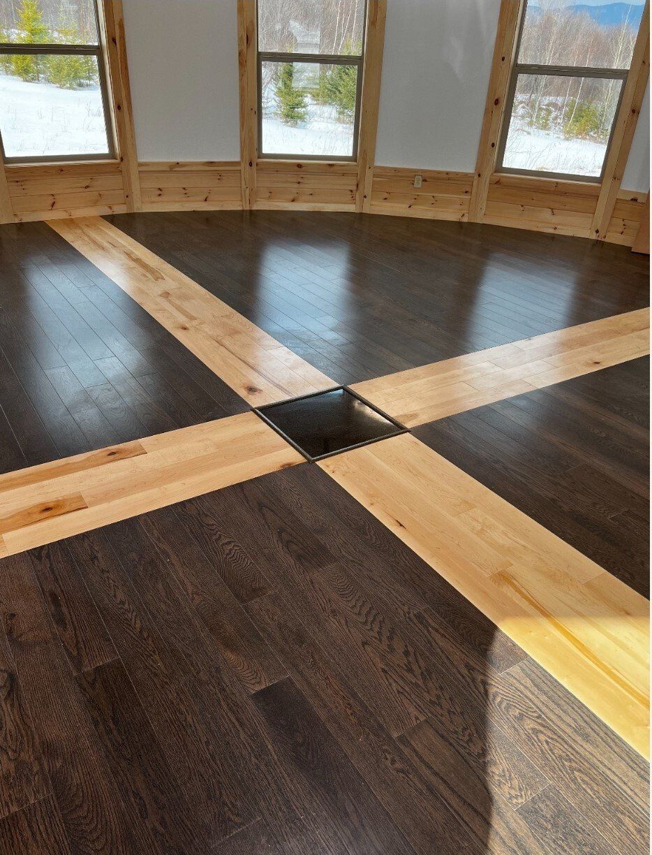 Wooden room floor with dark planks and light beams crossing to a central square near windows.