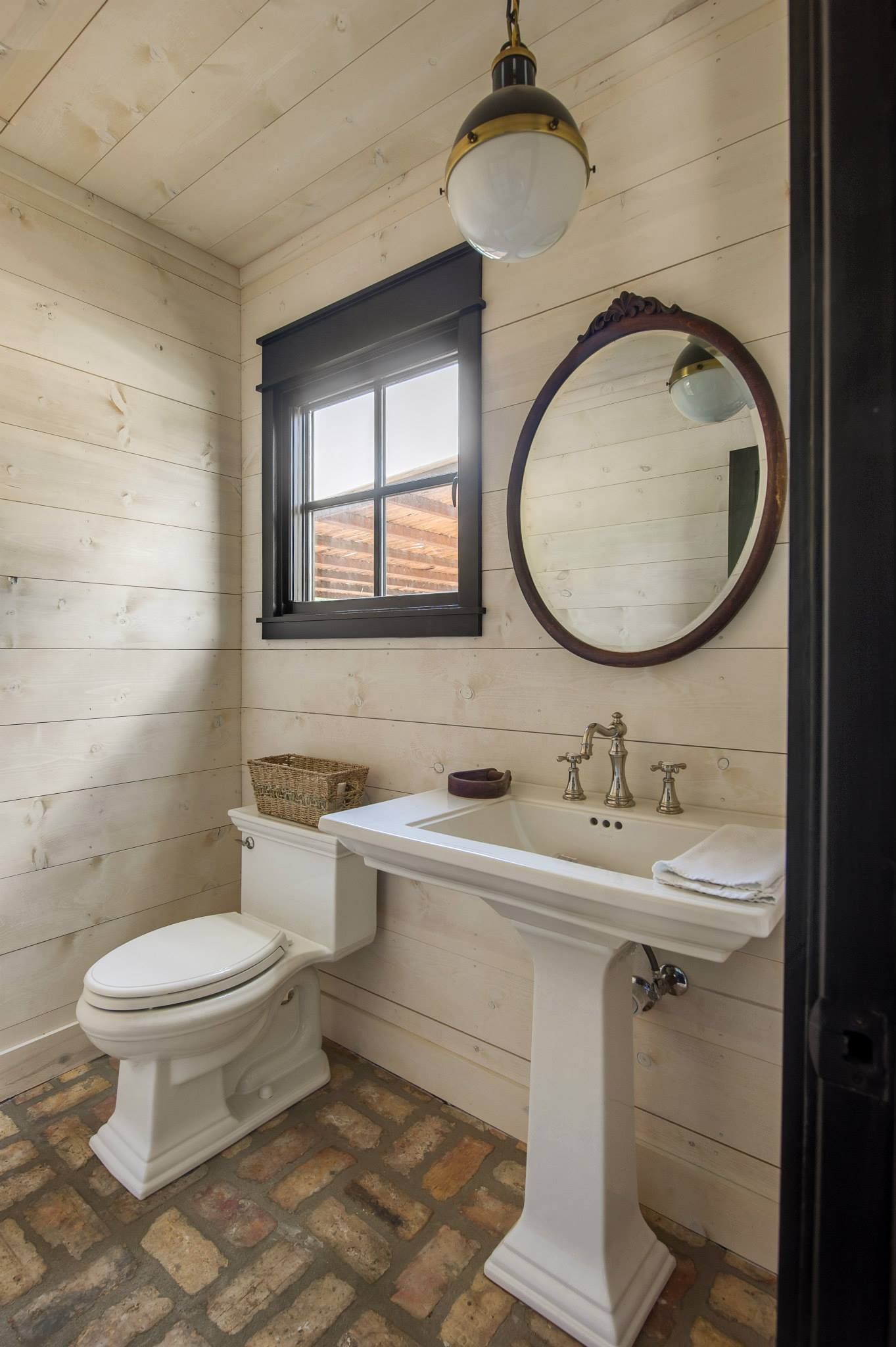 Small rustic bathroom with toilet, pedestal sink, oval mirror, window, and hanging light.