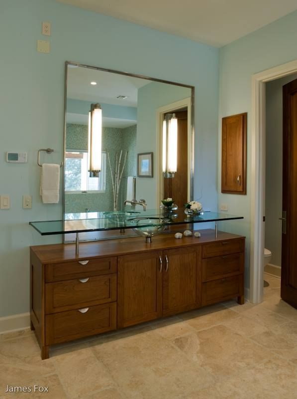 Bathroom vanity with glass countertop, mirror, and light blue walls beside an open doorway.
