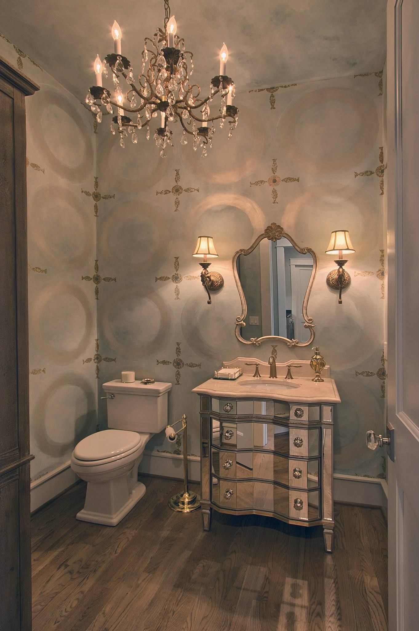 Elegant vintage bathroom with chandelier, ornate vanity, toilet, and mirrored wall sconces.