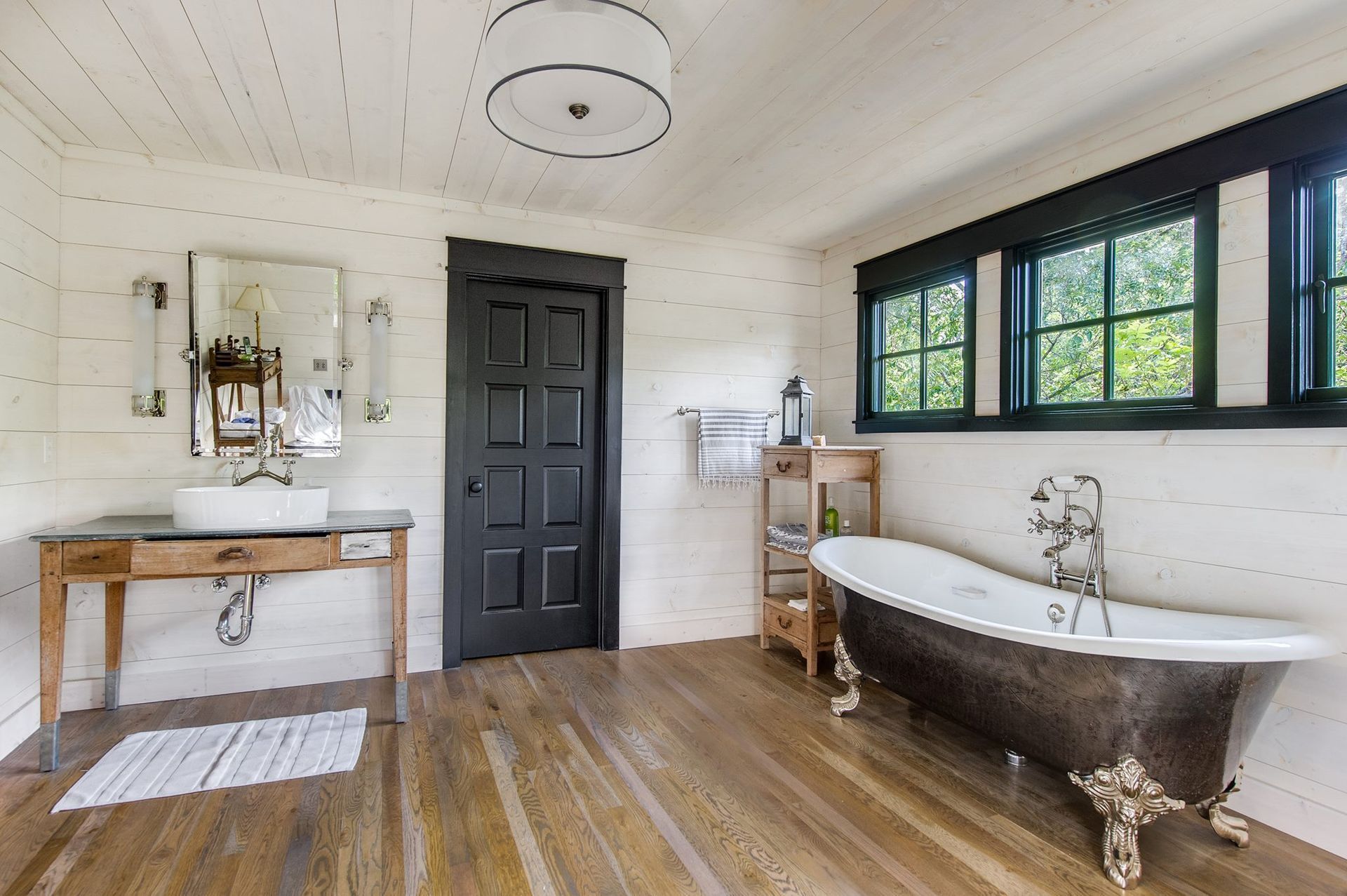 Bright bathroom with black door, wood vanity, freestanding tub, and large window.