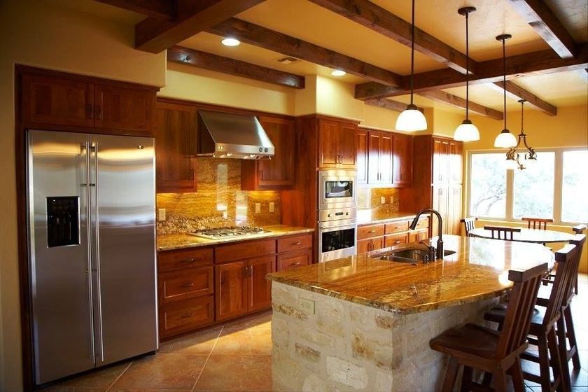 Warm kitchen with wood cabinets, stainless steel appliances, stone island, and bar stools by a window.