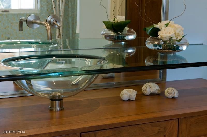 Glass bathroom sink with chrome faucet on a wooden vanity, beside a matching glass shelf and decor