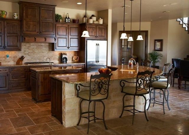 Warm kitchen with wood cabinets, stone island, pendant lights, and metal barstools on a tiled floor